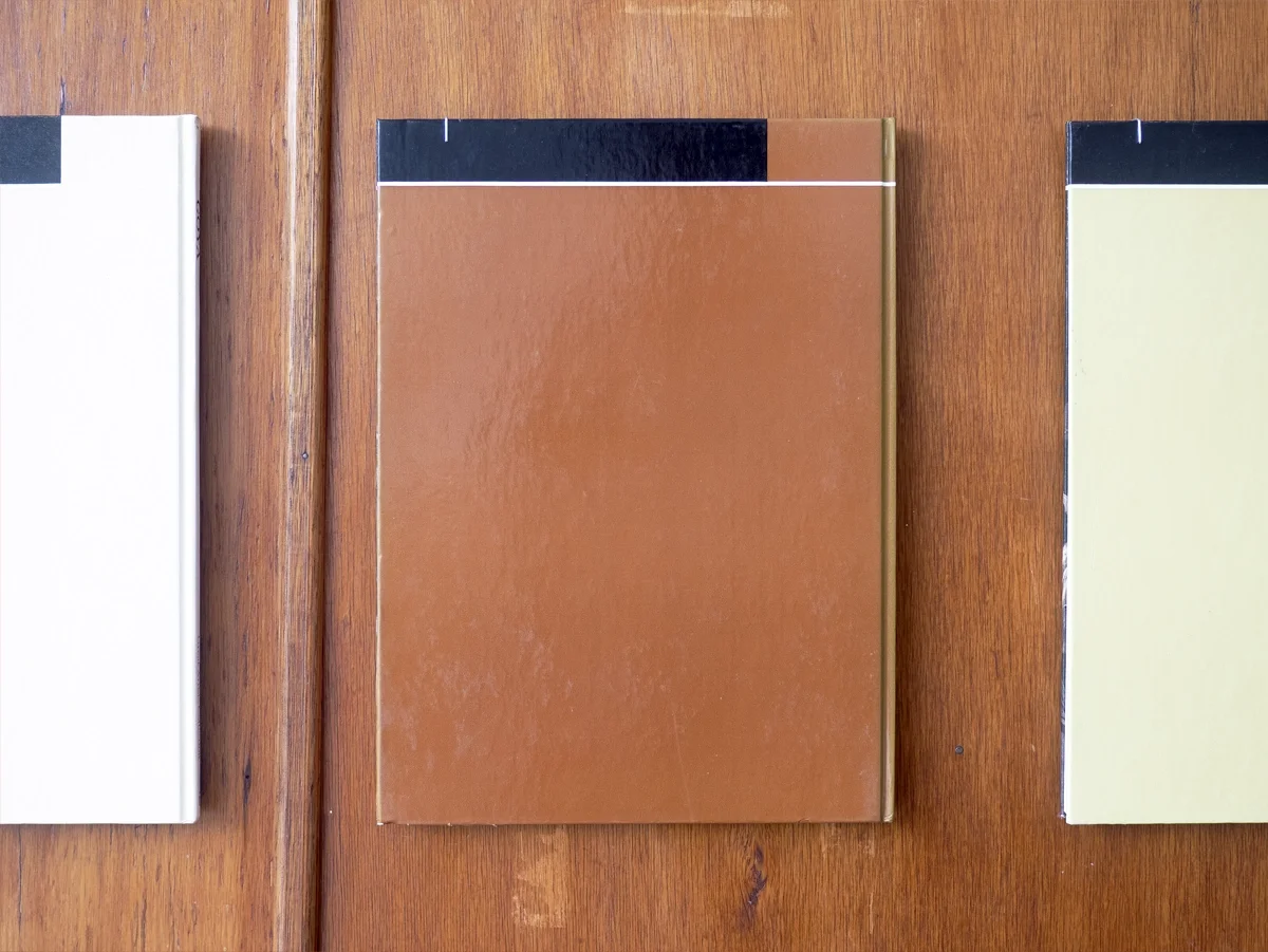 Three books with plain covers arranged on a wooden surface, with a brown book in the center and a white book partially visible on each side by artist Thomas Pendergast.