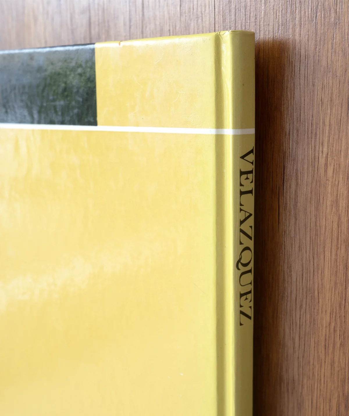 Close-up of a yellow book with partial text "VELAZQUEZ" on the spine, resting on a wooden surface. Artwork by artist Thomas Pendergast.