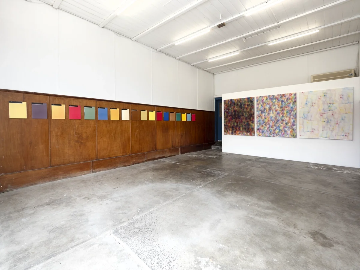An art gallery with colorful abstract artwork by artist Thomas Pendergast on the wall and multicolored paper squares displayed on a wooden panel.