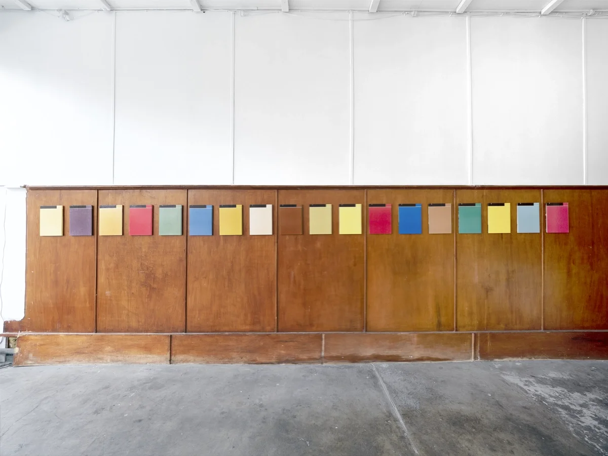 Colorful notebooks or folders by artist Thomas Pendergast displayed on a wooden wall shelf in an indoor setting.
