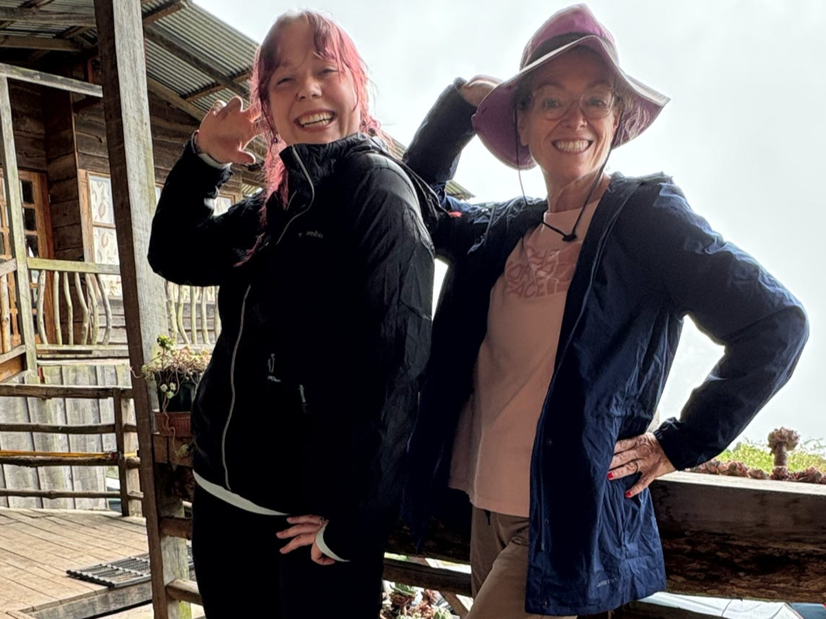 The Hygiene Ladies co-founders, Hailee and Kim, standing outdoors at a wooden building, smiling at the camera. One woman has pink hair and is wearing a black jacket, the other woman has glasses, a pink hat, and a blue jacket.