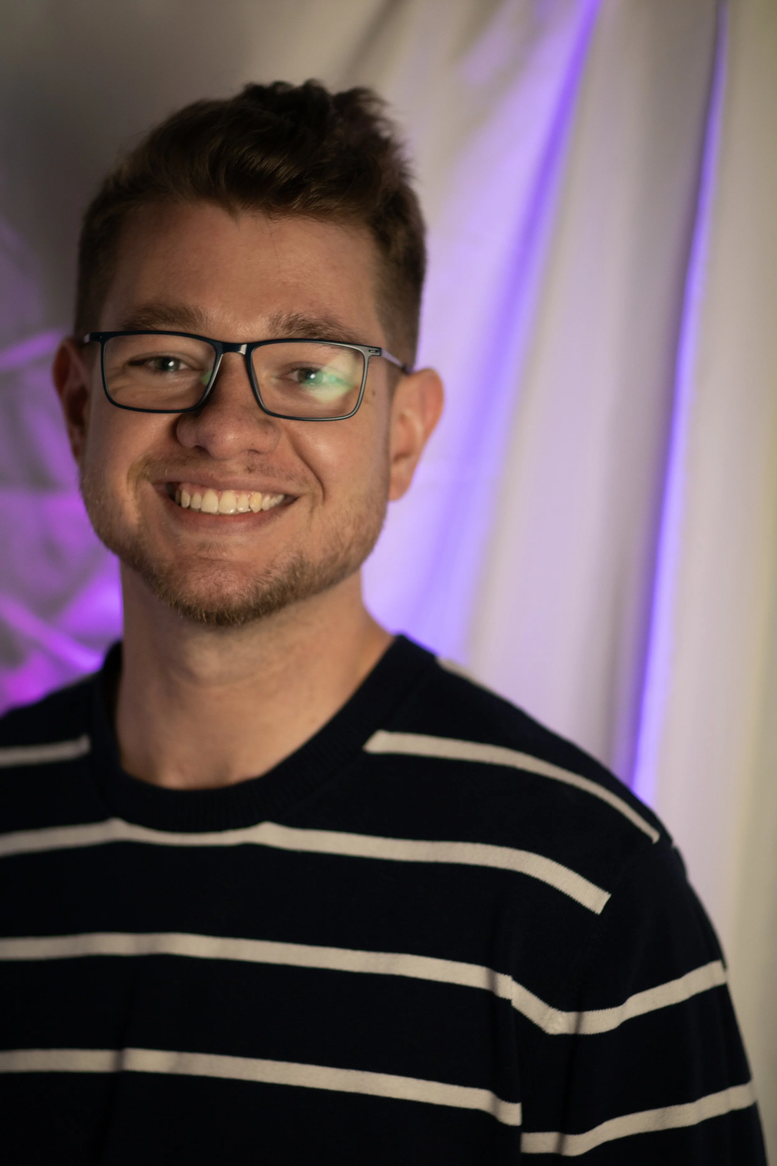 A young Hygiene Ladies board member with glasses smiling at the camera, wearing a black and white striped shirt, with a purple light in the background.