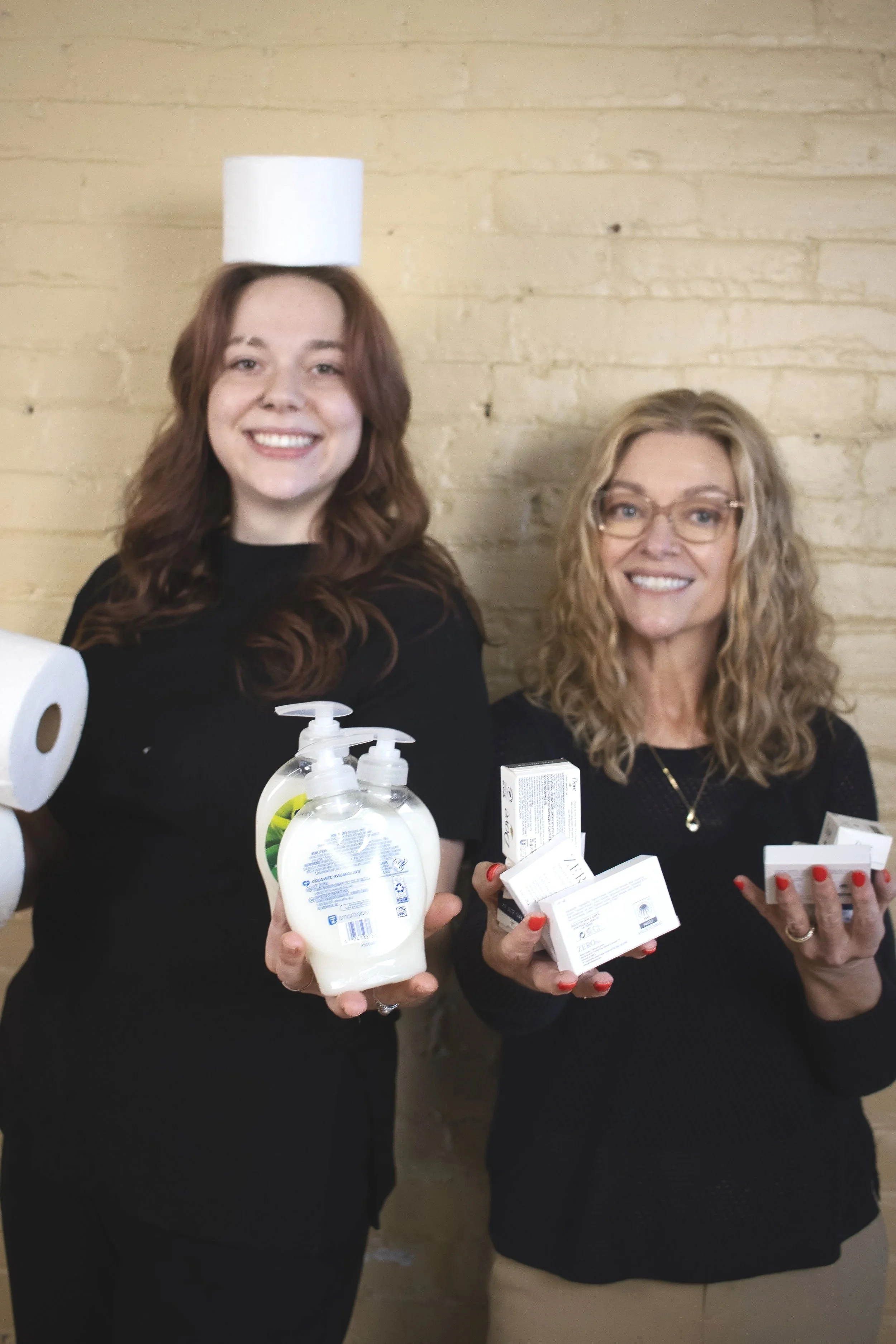 Two women smiling and holding various personal hygiene products, including hand sanitizer and tissue boxes, standing in front of a yellow brick wall.