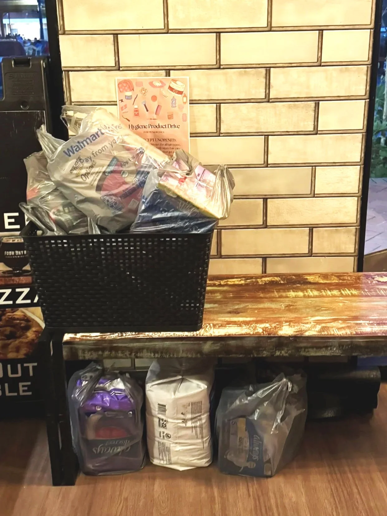 A wooden bench with three bags and a basket of wellness products on top. The bags are plastic and contain various donated items. A brick wall is in the background.