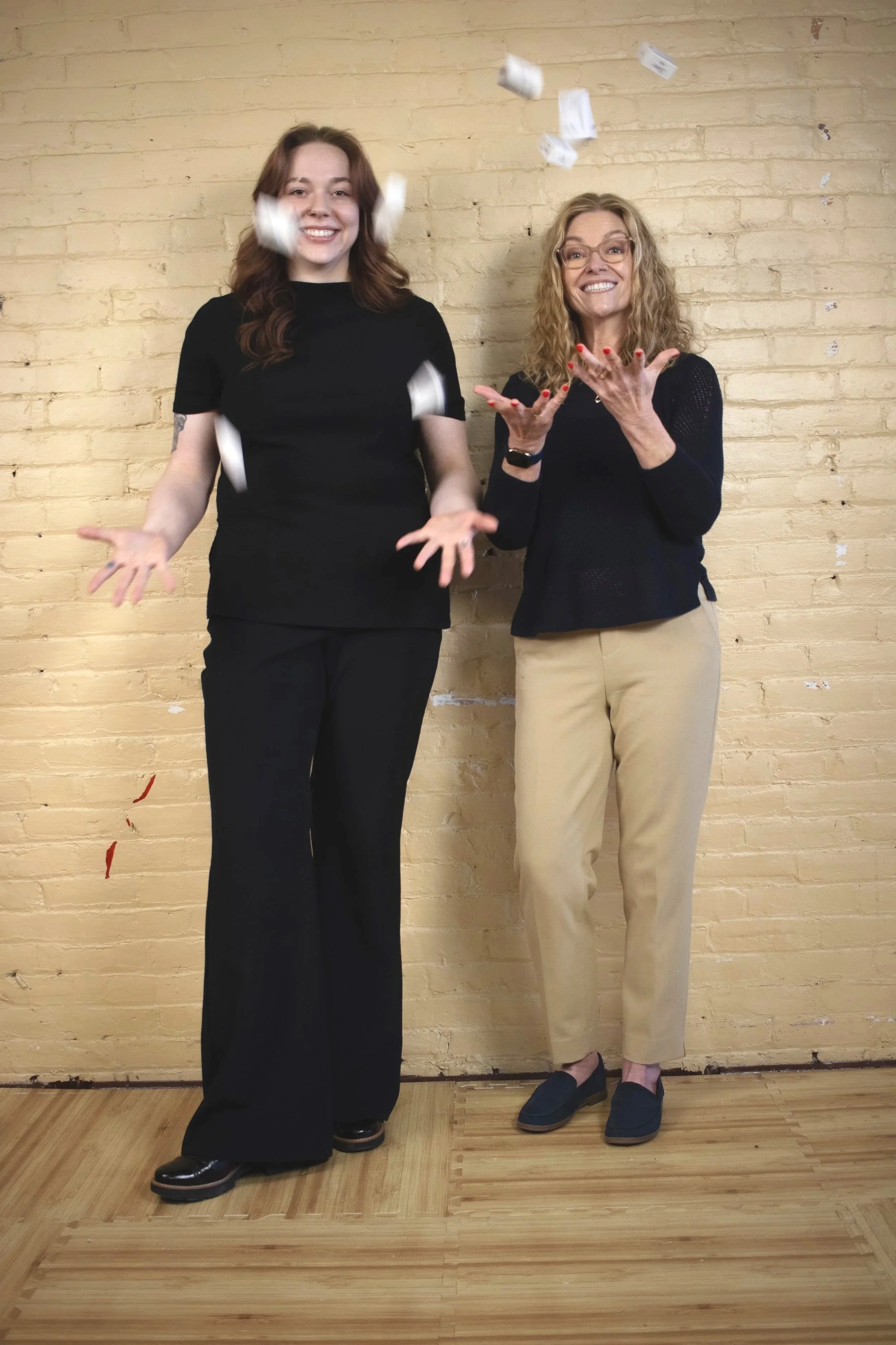 The two co-founders of The Hygiene Ladies standing in front of a yellow brick wall, smiling, tossing rolls of toilet paper into the air to celebrate the founding of Muncie's hygiene pantry.