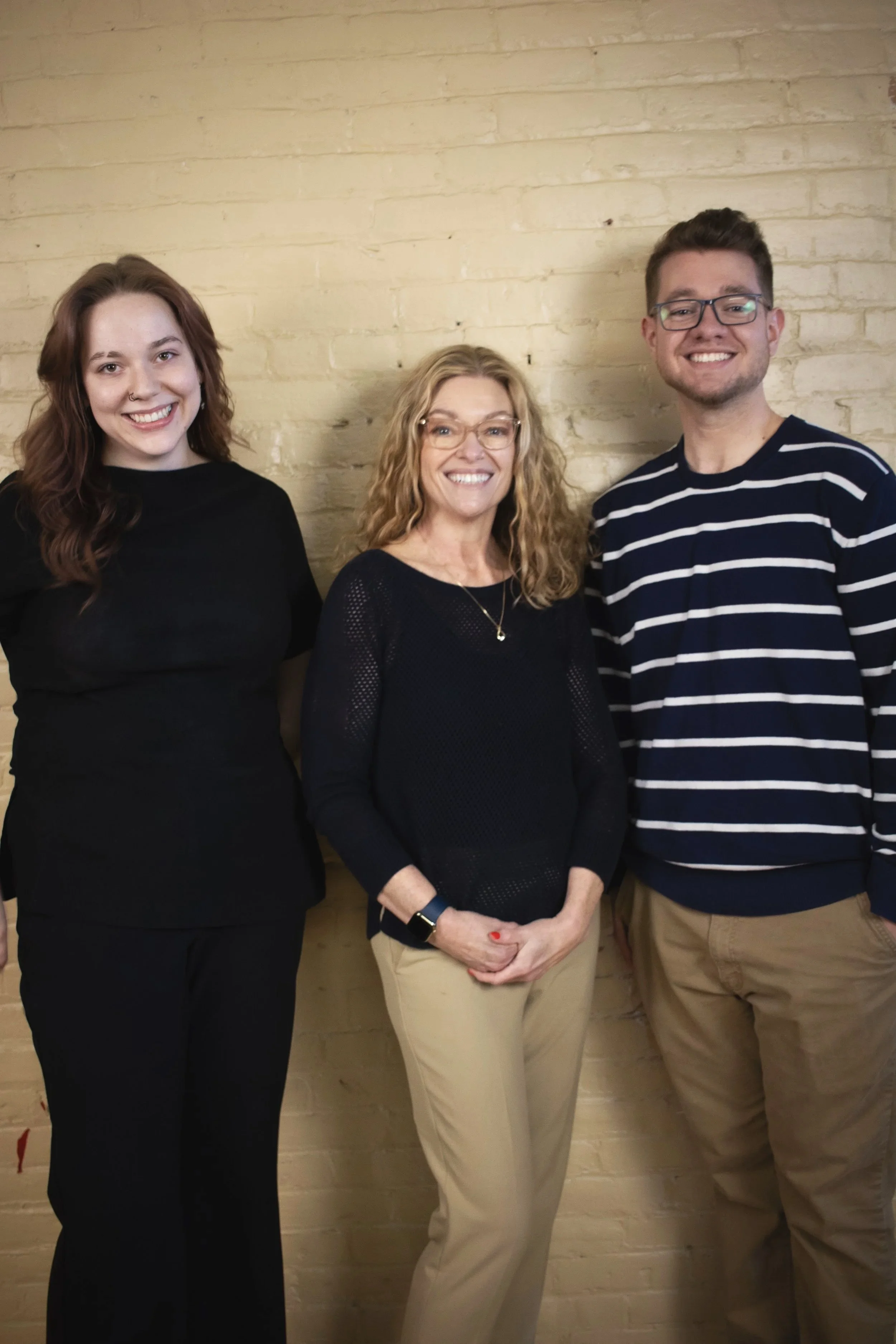 Three people standing in front of a beige brick wall, smiling at the camera. The person on the left is a woman with long, wavy, auburn hair wearing a black top and black pants. The person in the middle is a woman with curly, blonde hair, wearing glasses, a black sweater, and beige pants. The person on the right is a man with short brown hair, glasses, a navy blue and white striped sweater, and beige pants.