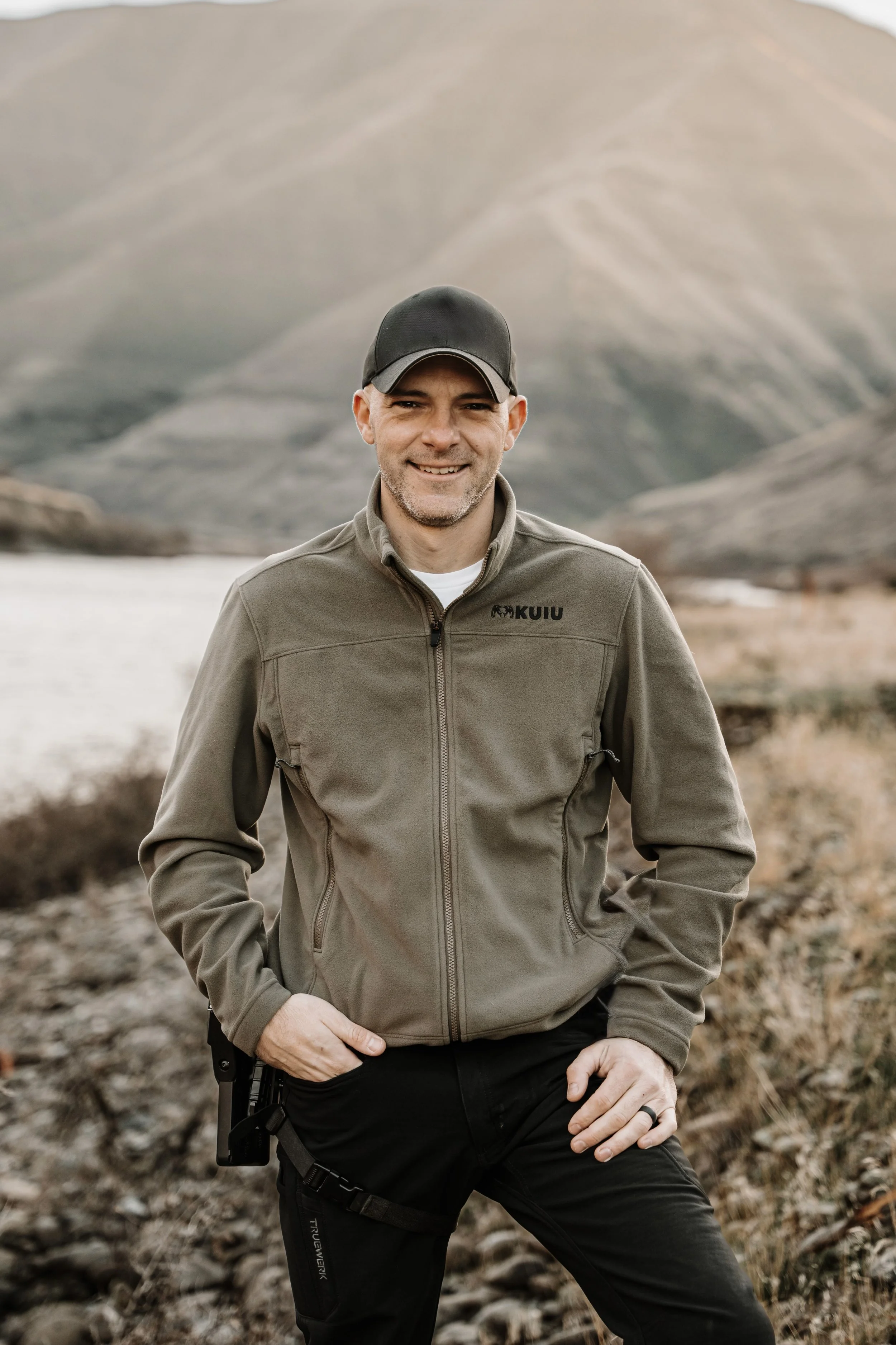 A man standing outdoors near a river and mountains, wearing a gray jacket and black cap, smiling at the camera.