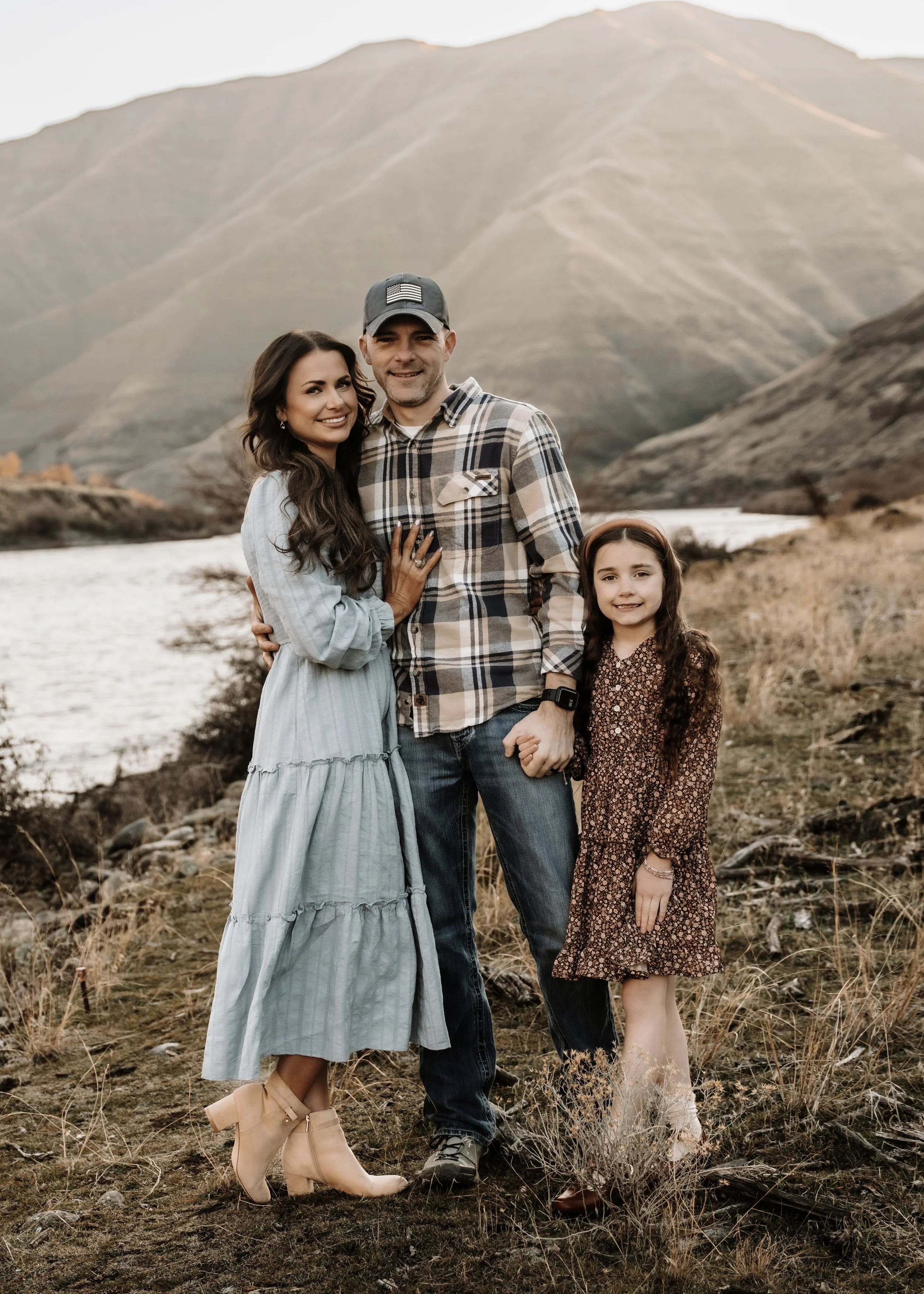 A family of three standing outdoors near a river with mountains in the background during sunset. The woman is wearing a blue dress and beige ankle boots, the man is in a plaid shirt and jeans, and the little girl is in a brown floral dress with tan shoes. They are smiling and holding hands.