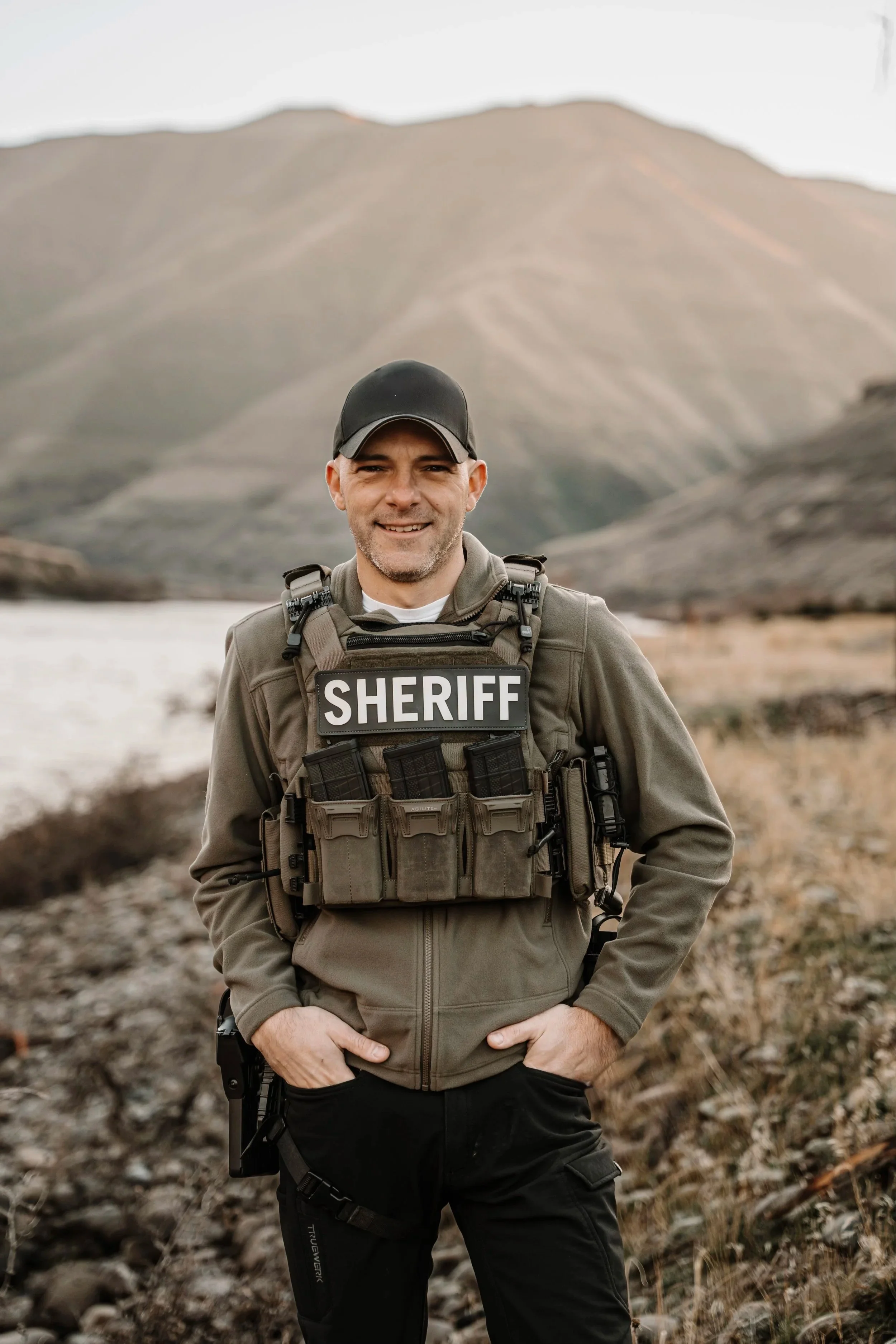 A male sheriff in uniform standing outdoors near a river with mountains in the background, smiling at the camera.