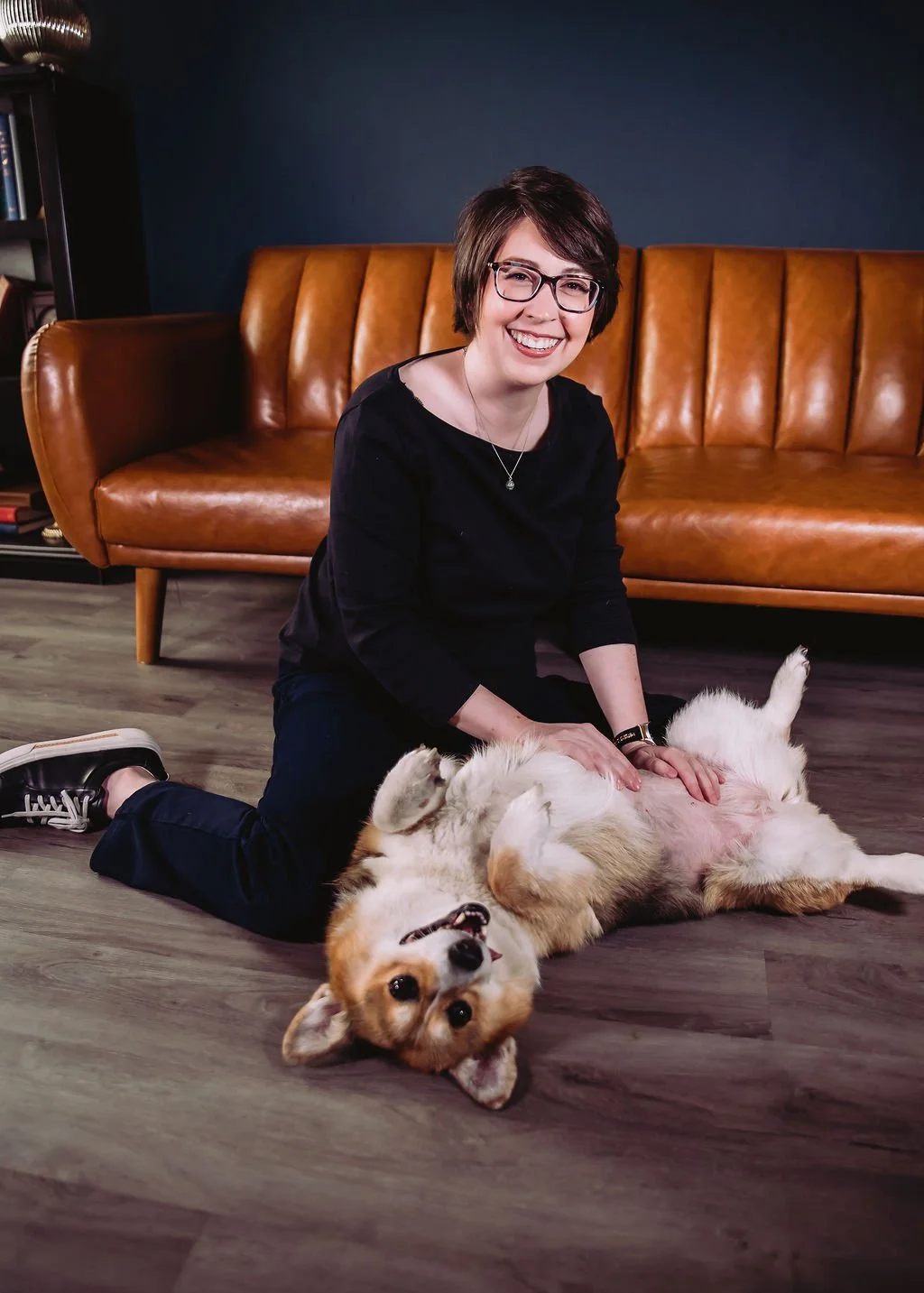 A woman with short brown hair, glasses, and a black top smiling while petting a corgi lying on the floor on its back. They are in a living room with a brown leather couch and a dark blue wall.