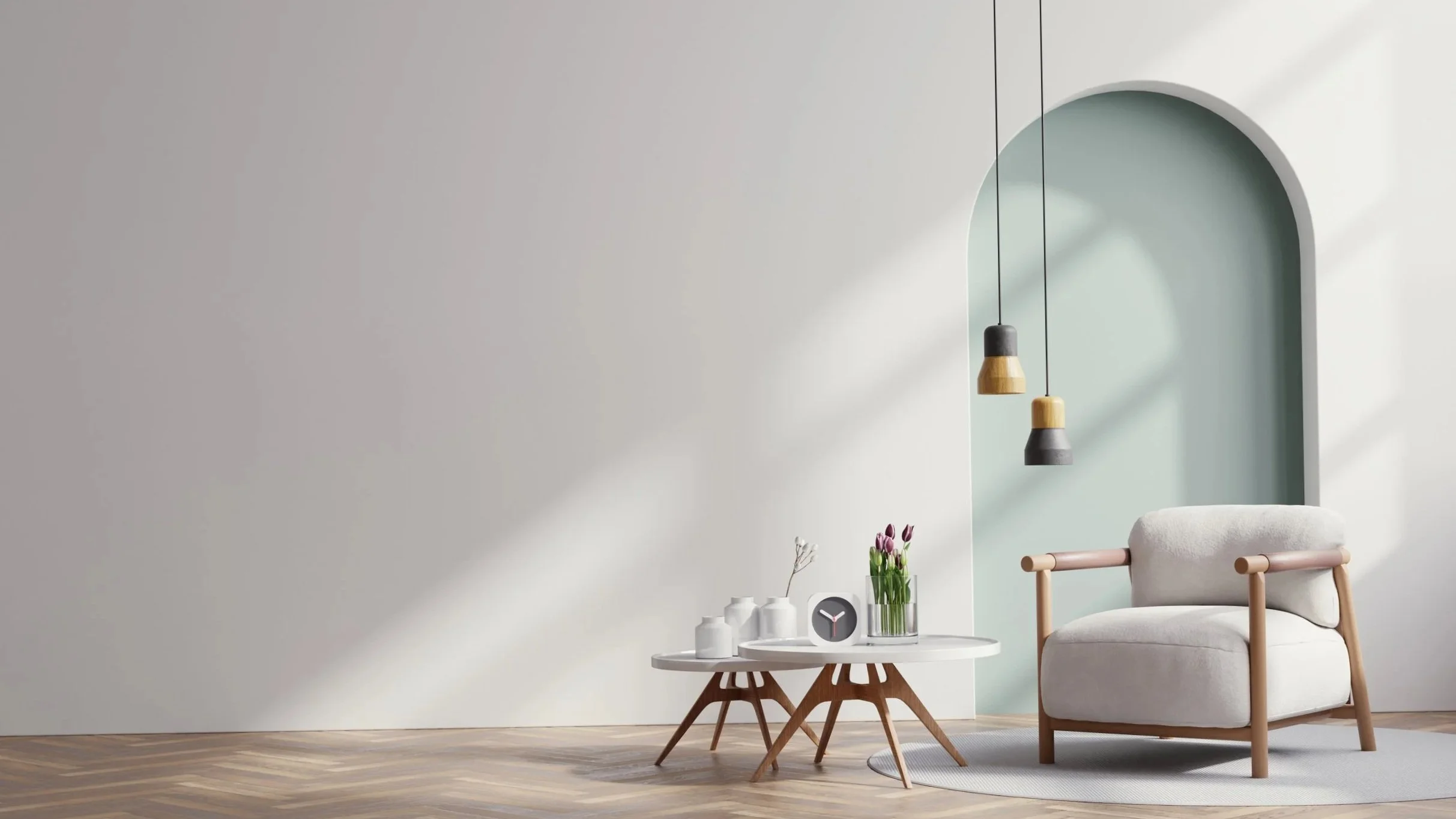 Minimalist living room with a white armchair with wooden arms, a small round white coffee table with wooden legs, a vase with tulips, a clock, and three wall-mounted pendant lights in black and beige hanging from the ceiling. Sunlight casts shadows on a white wall with an arched alcove painted light green.
