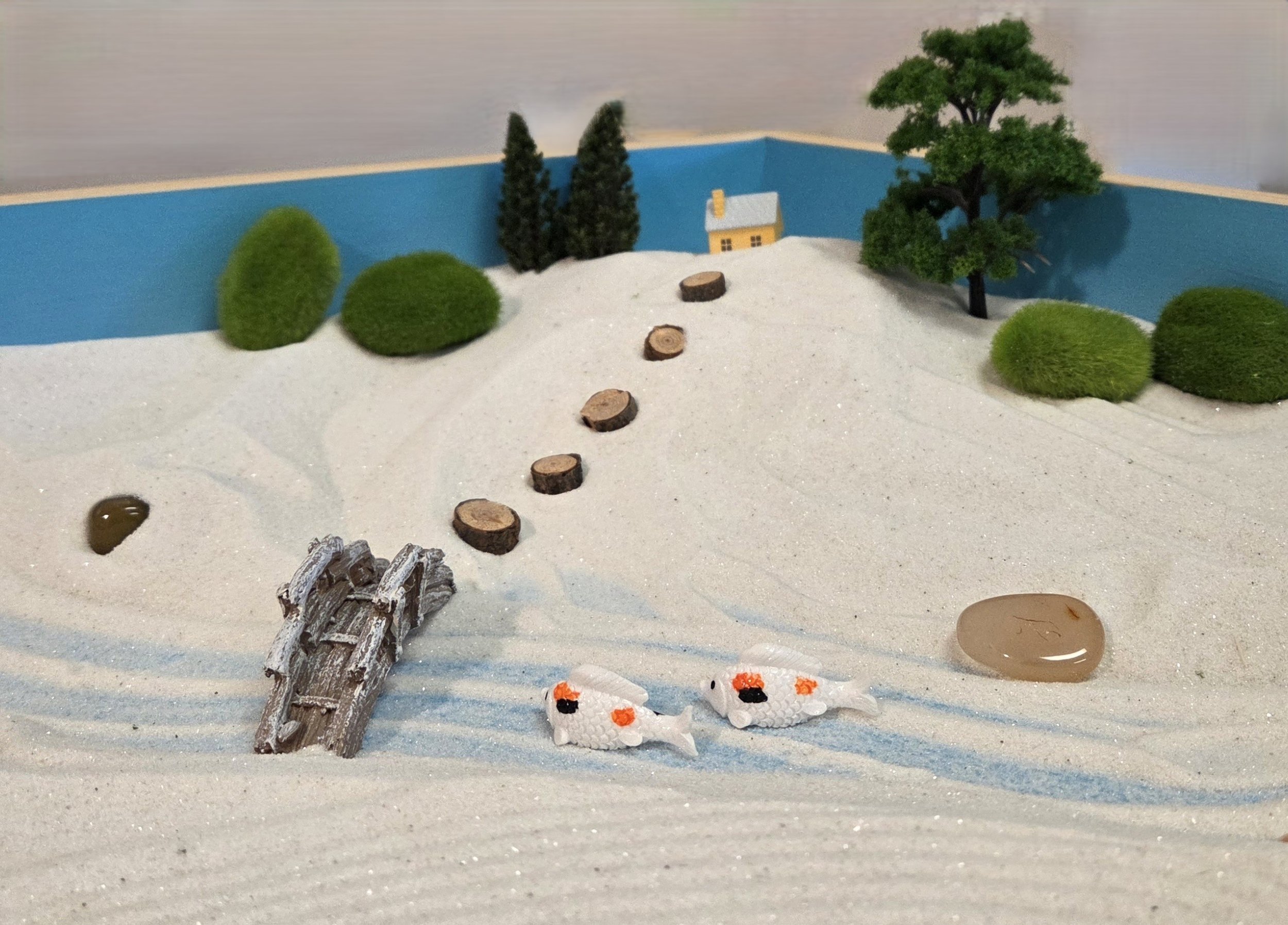 Miniature winter scene with snow-covered ground, small trees, a tiny house, a wooden bridge, and two small fish in the snow.