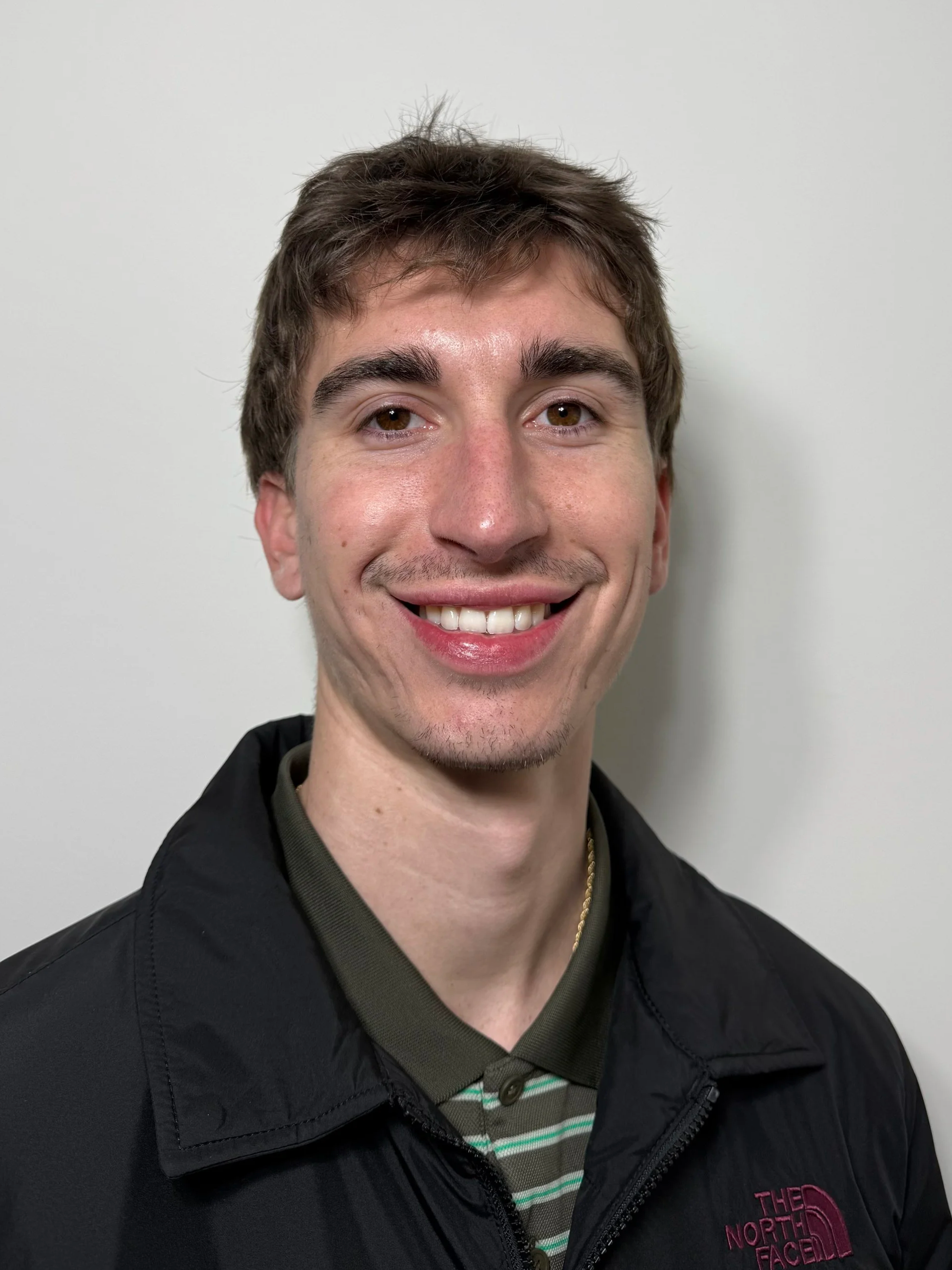 A young man with brown hair, light skin, and a big smile, wearing a striped shirt and a black North Face jacket, standing against a plain white wall.