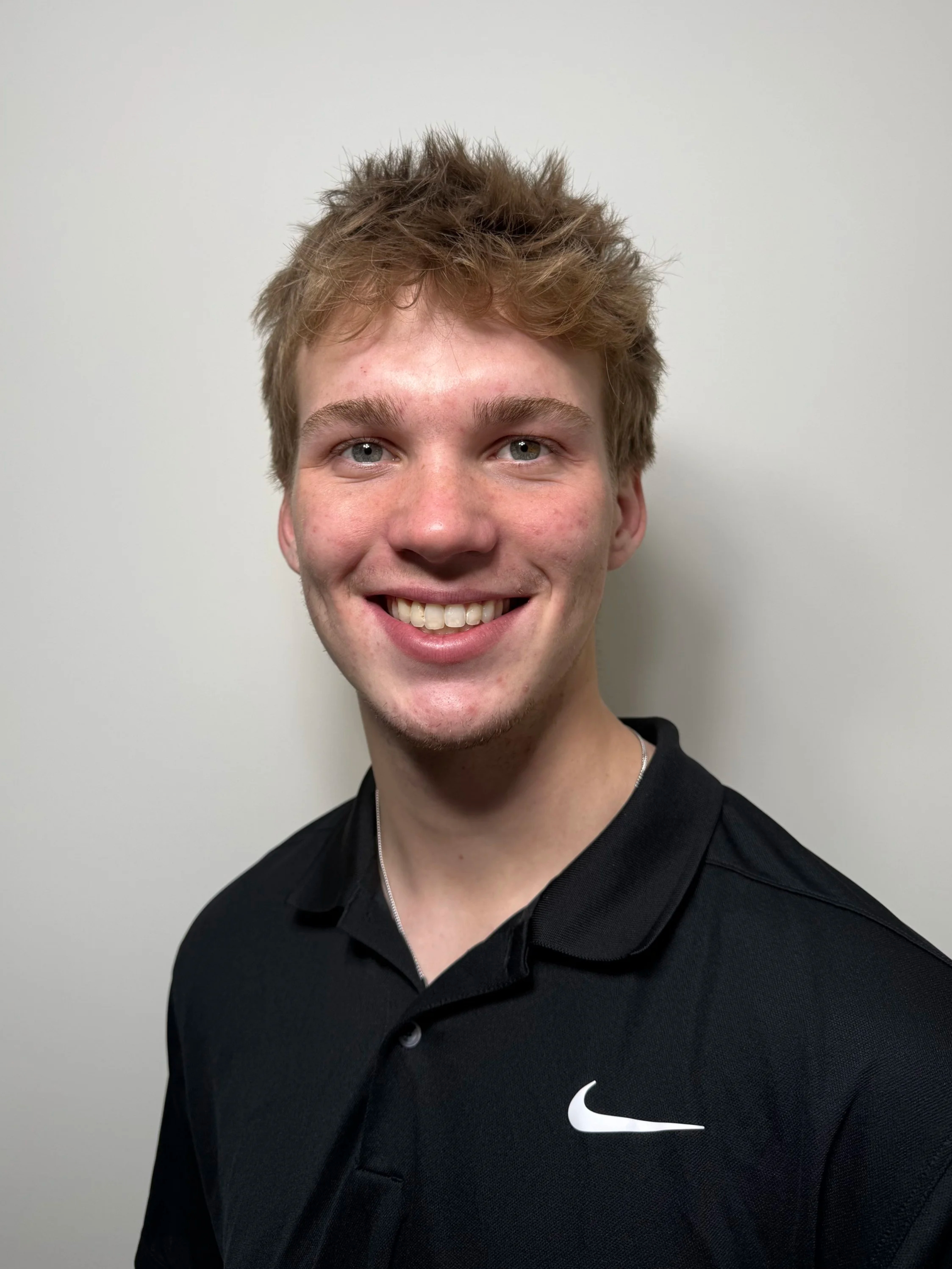 A young man with light brown, tousled hair and blue eyes smiling, wearing a black Nike polo shirt, standing against a plain off-white wall.