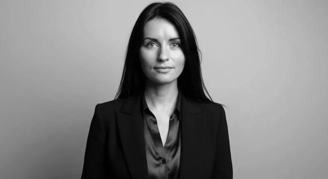 Black and white portrait of a woman with long dark hair, wearing a dark blazer and satin blouse, standing against a plain background.