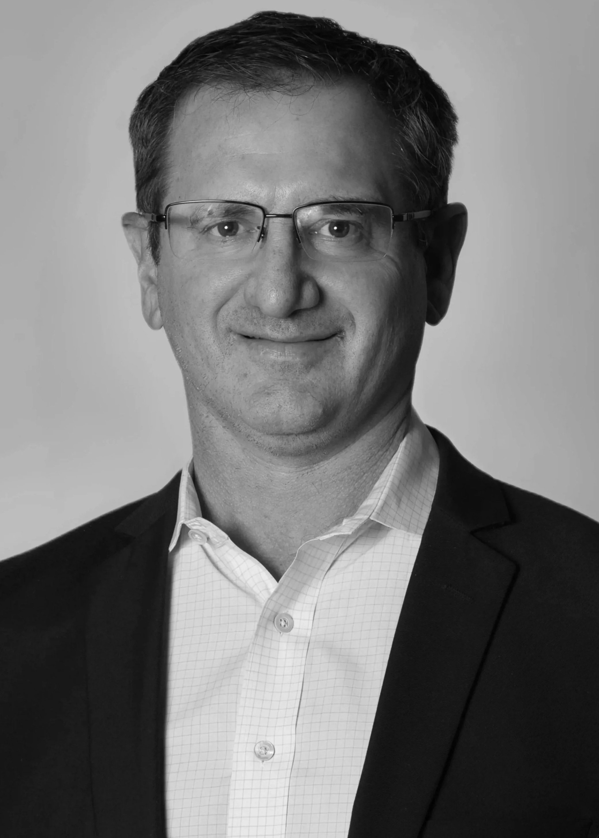Black and white professional headshot of a man wearing glasses, a light-colored checkered shirt, and a dark blazer, smiling.