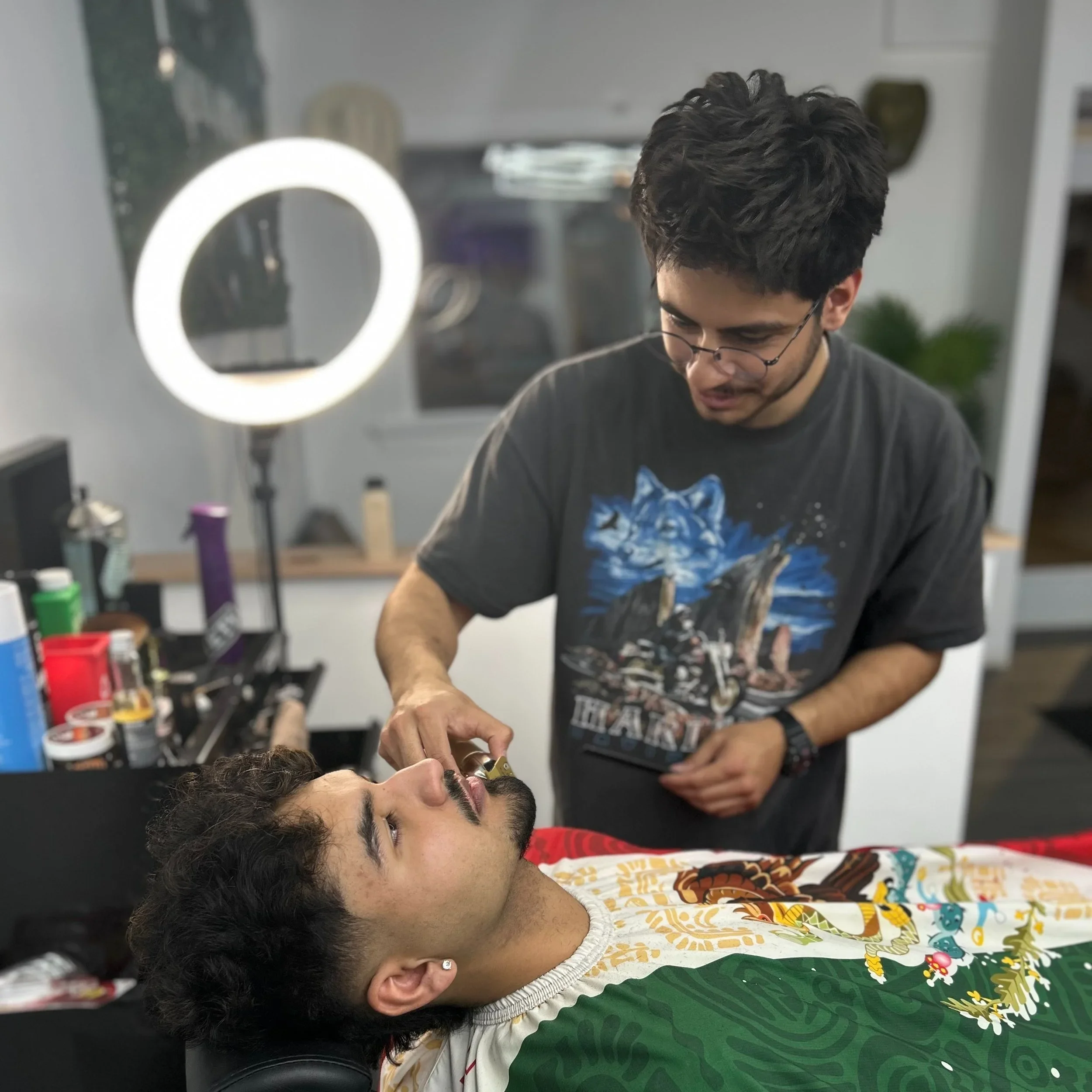 A young man with dark curly hair and a beard is lying down with his head on a black surface. Another young man wearing glasses and a dark graphic T-shirt is shaving his face with an electric razor. The background includes a ring light, shelves with various grooming supplies, and a mirror.