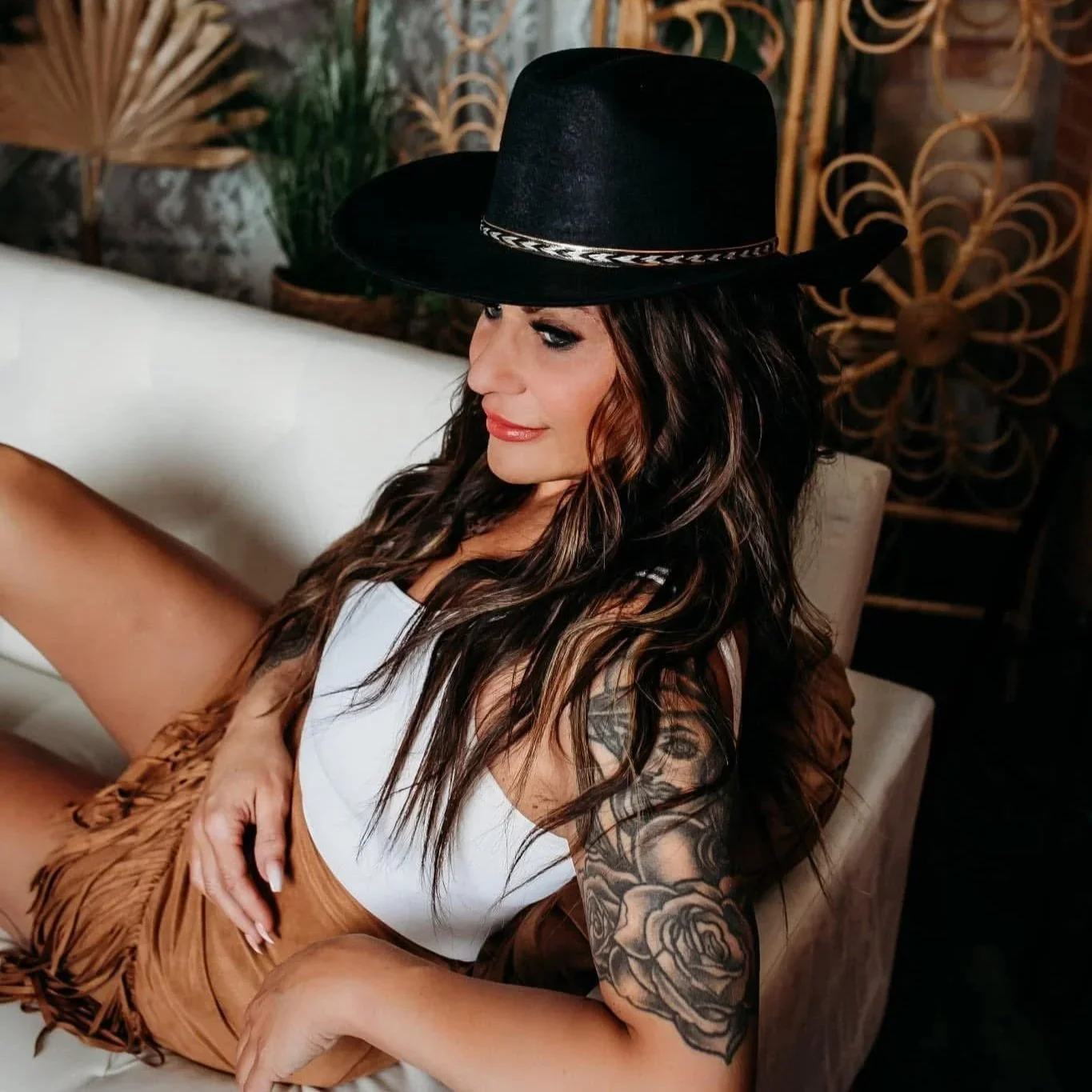 A woman with long wavy hair wearing a large black hat, white tank top, and tan shorts, sitting on a white couch with a tattooed arm. In the background, there are decorative plants and intricate golden metal latticework.
