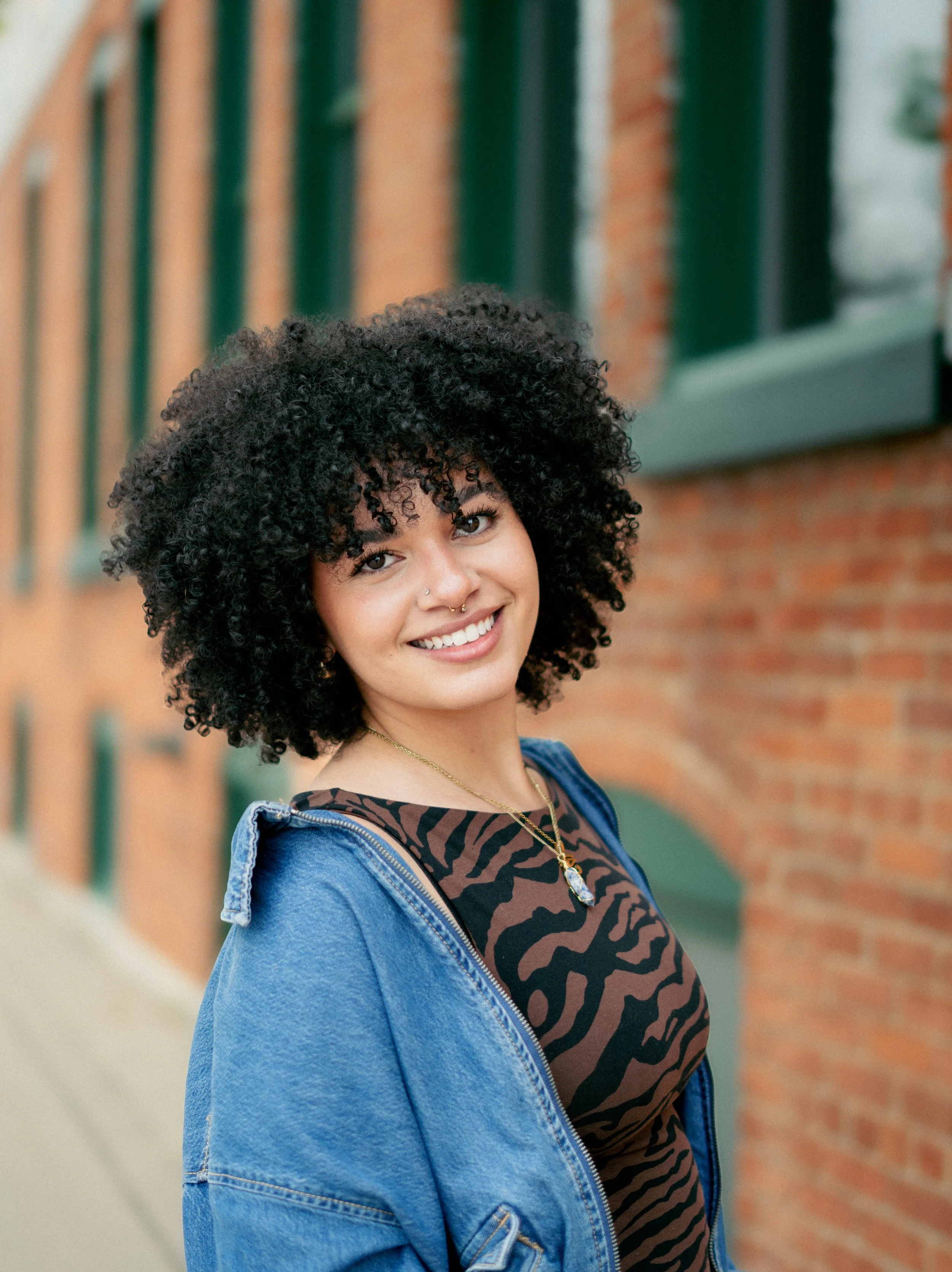A young woman with curly black hair, smiling and wearing a nose ring and a statement necklace makes eye contact with the camera while standing outside in front of a brick building with green window frames, wearing a denim jacket over a brown and black animal print top.