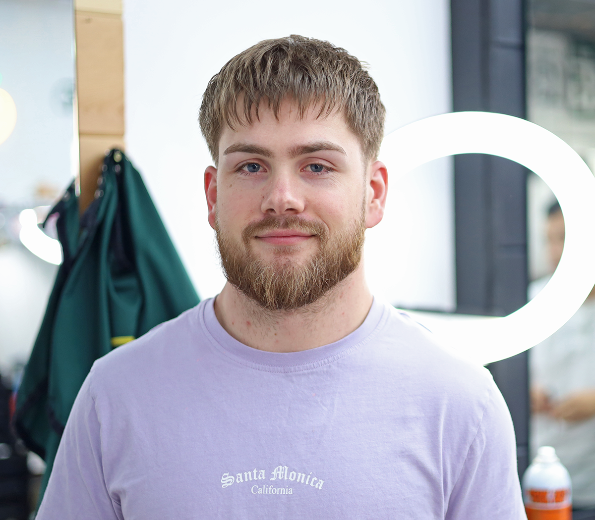 Portrait of a young man with a beard, wearing a light purple T-shirt that says Santa Monica California, standing in a room with a mirror illuminated by LED lights.