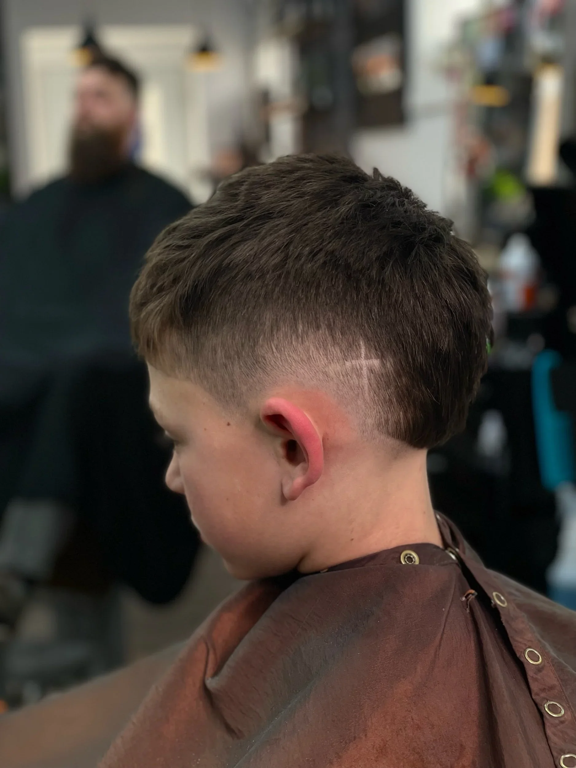 A young man with a freshly cut hairstyle, featuring a fade on the sides and back, is sitting in a barbershop. The barber in the background is blurred, focusing on the client's haircut.