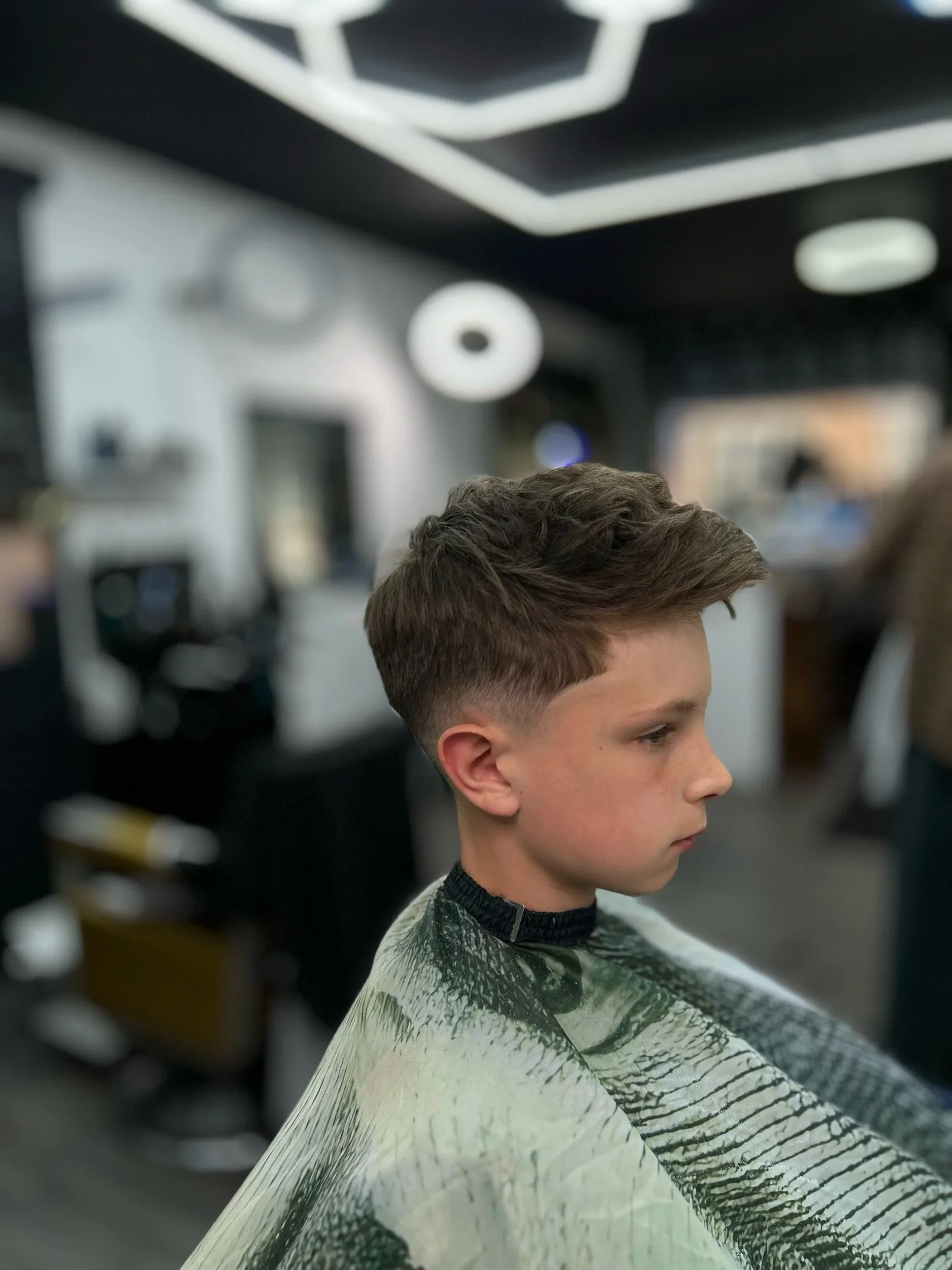 A young boy with a freshly styled haircut sitting in a barber shop, wearing a barber cape, with a blurred background of the shop interior.