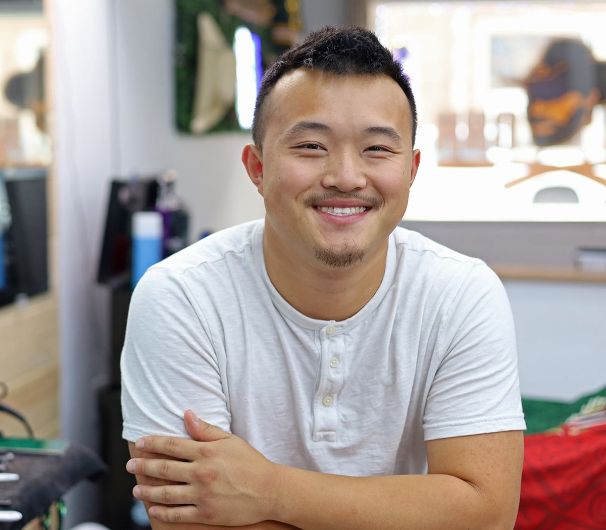 A smiling young man with short black hair, a slight beard, and a mustache, wearing a white shirt, sitting indoors with a blurred background including a guitar and various objects.