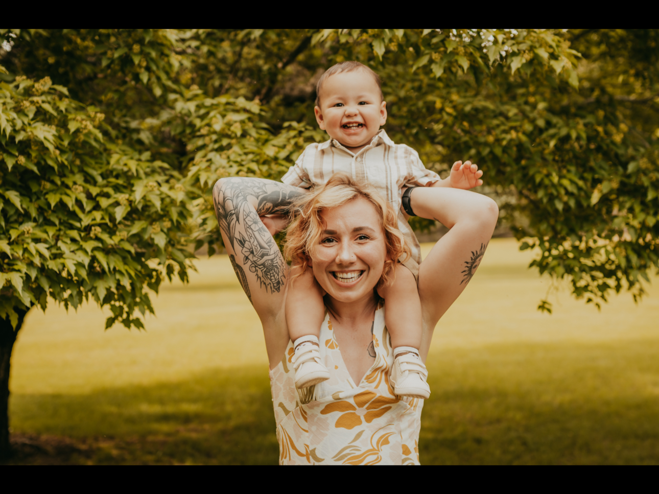 A smiling woman with blonde hair and tattoos on her arms carrying a happy toddler on her shoulders in a park with trees.