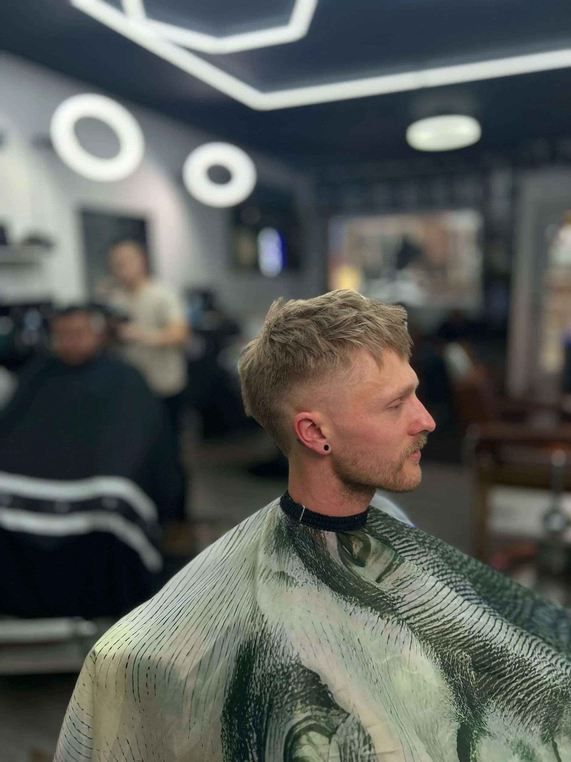 Young man with short, textured haircut sitting in a barbershop, wearing a barber cape with a swirling pattern, in a modern interior with other patrons and a barber in the background.