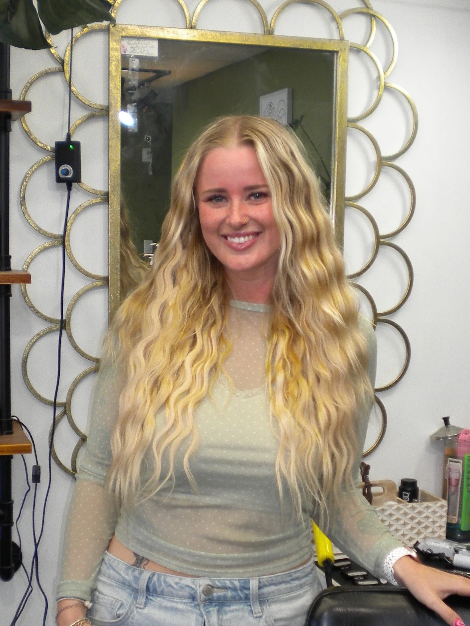 Smiling woman with blonde hair, yellow cropped sweater, and tattoos, standing in a salon.