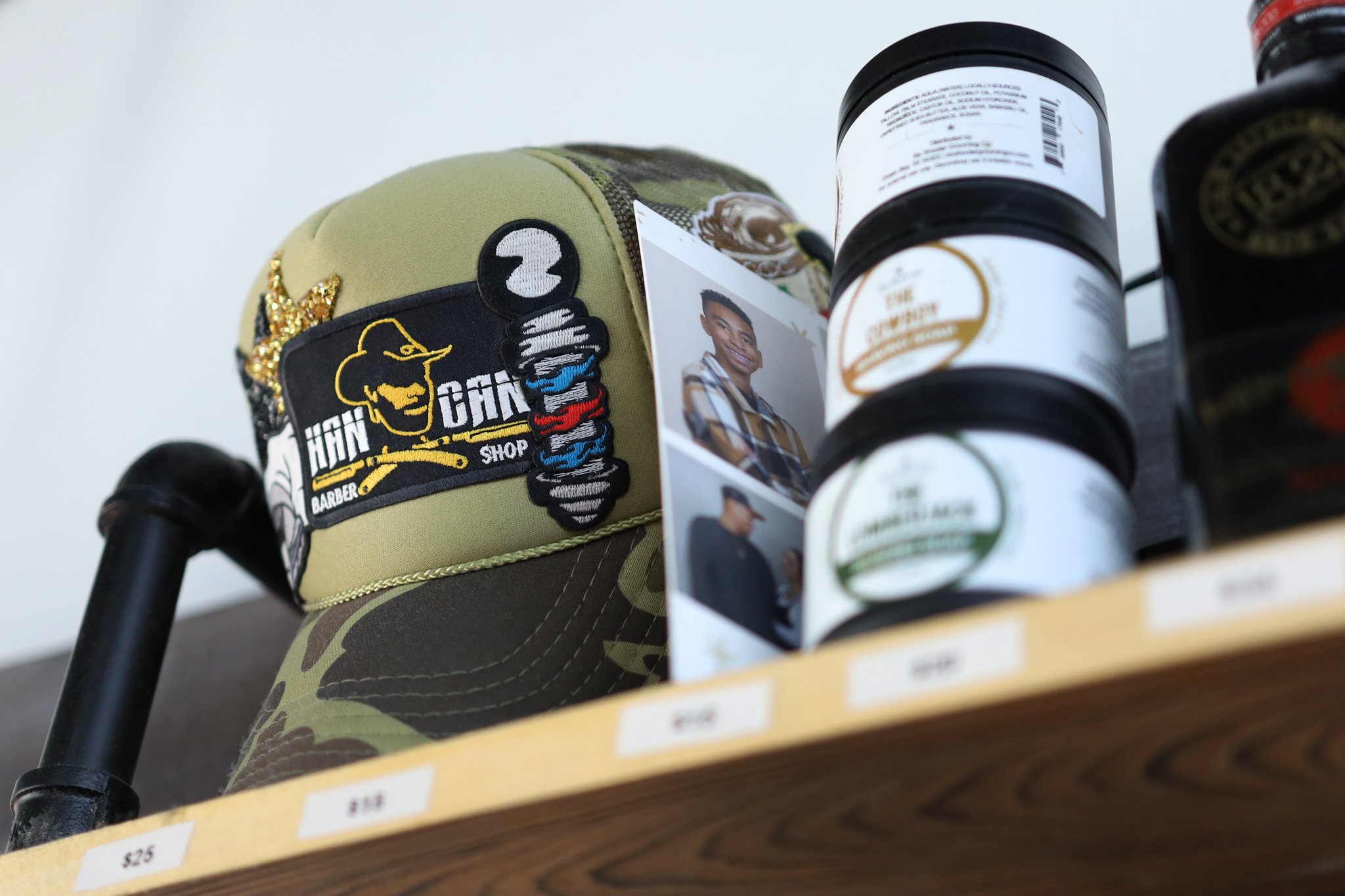 Close-up of a baseball cap with embroidered patches, including a barber pole and a face, displayed on a store shelf alongside bottles of grooming products.