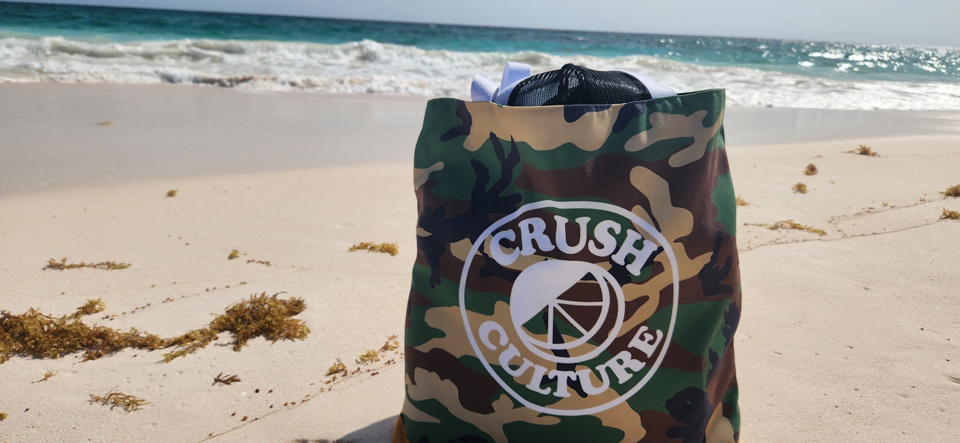 A camouflage tote bag with 'Crush Culture' logo on a sandy beach near the ocean, with seaweed scattered on the sand and waves in the background.