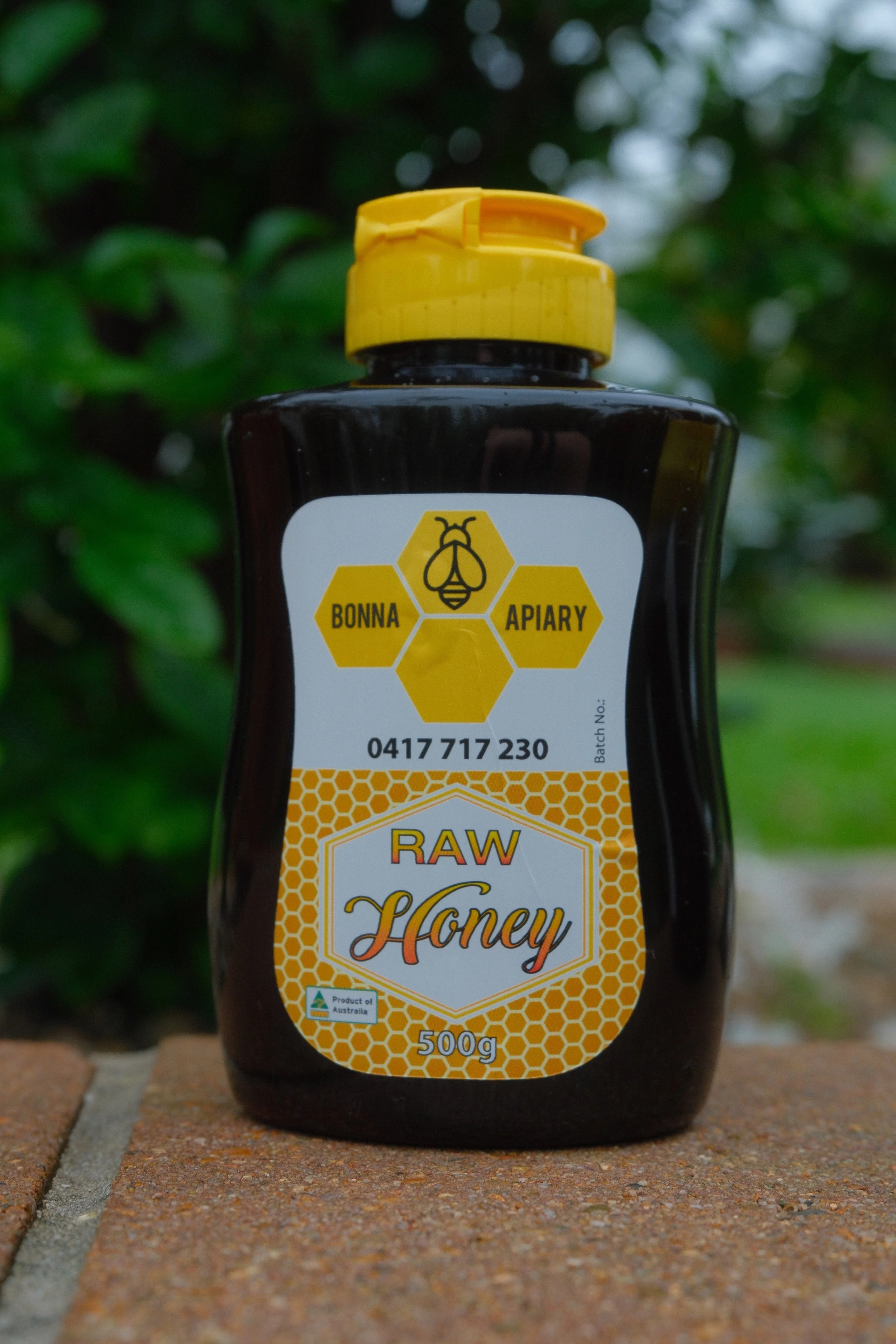 A jar of raw honey with a yellow flip cap, placed on a brick surface outdoors with green foliage in the background.