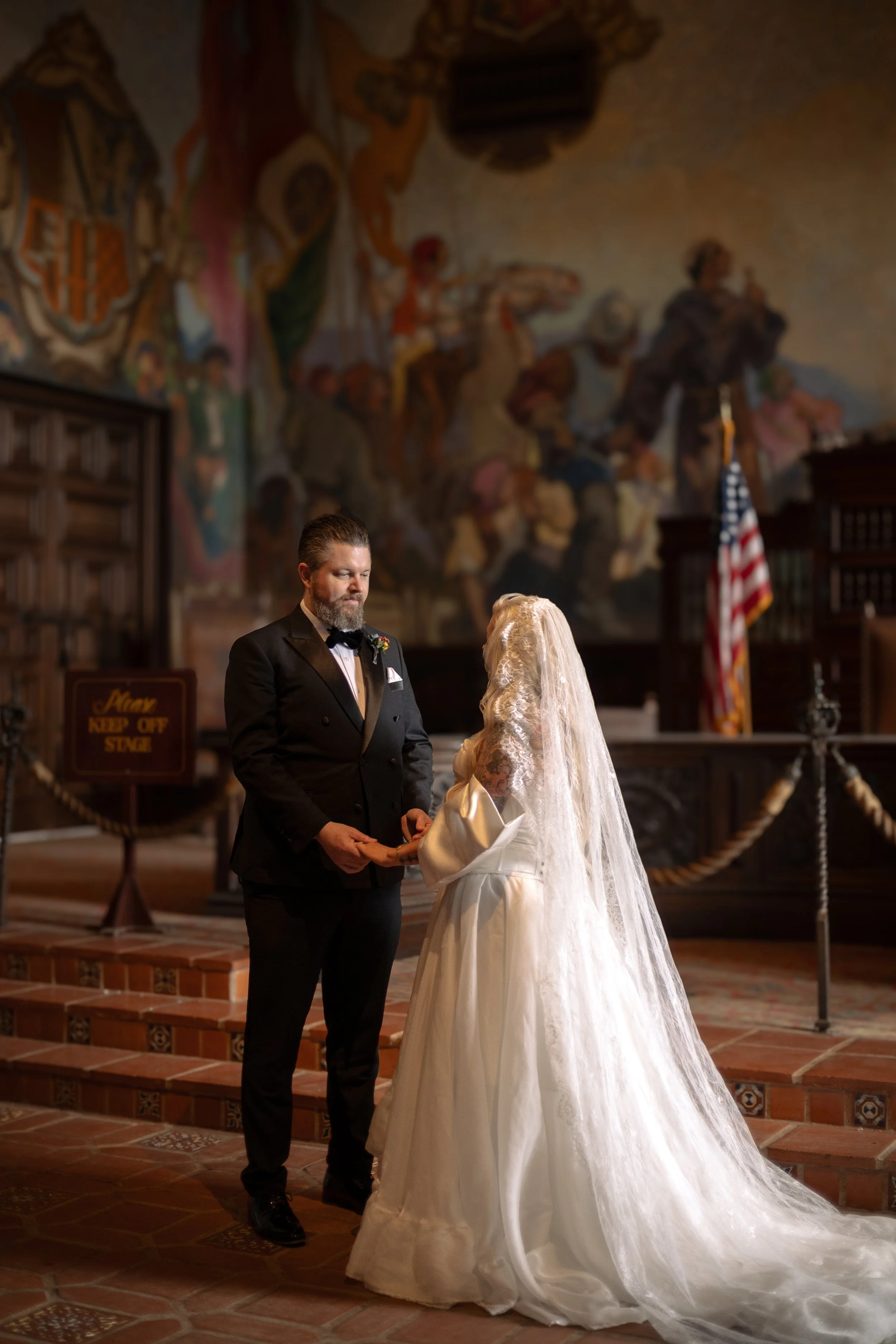 A couple getting married in a courthouse, with the groom in a black tuxedo and the bride in a white wedding dress with a long veil, standing on steps in front of an altar.