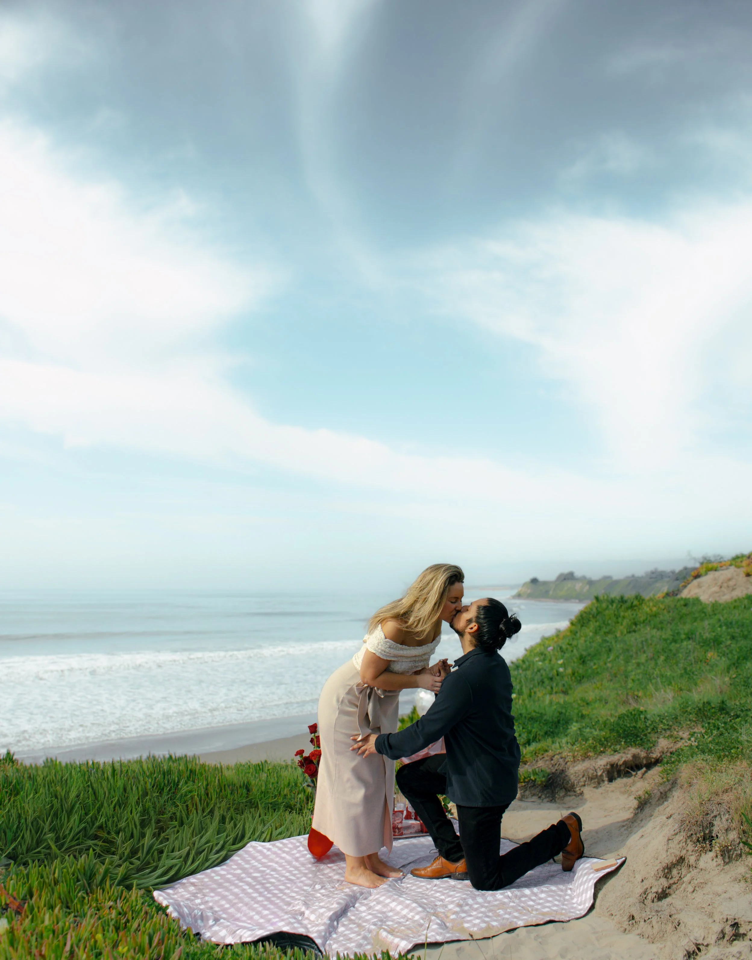 A man kneeling on one knee in front of a woman on a beach, proposing marriage. They are holding hands and kissing, with a blanket on the sand and a scenic Santa Barbara cliff and ocean background.