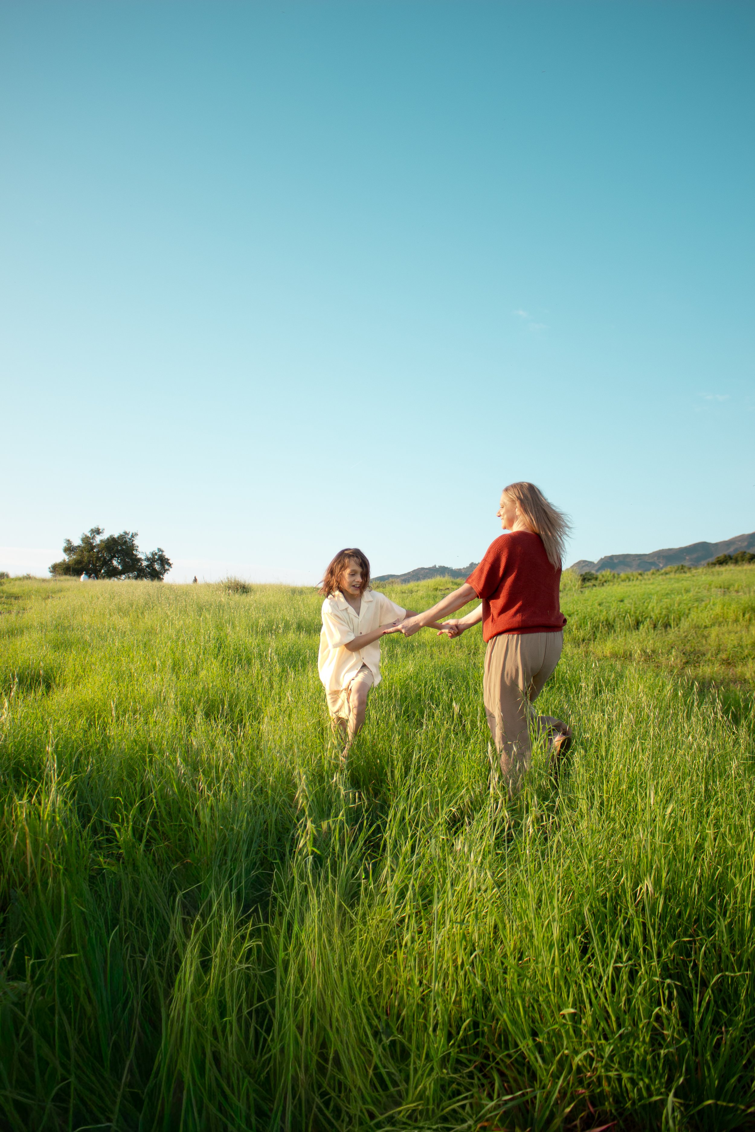 A woman and a young boy playing and holding hands in a lush green field under a clear blue sky.