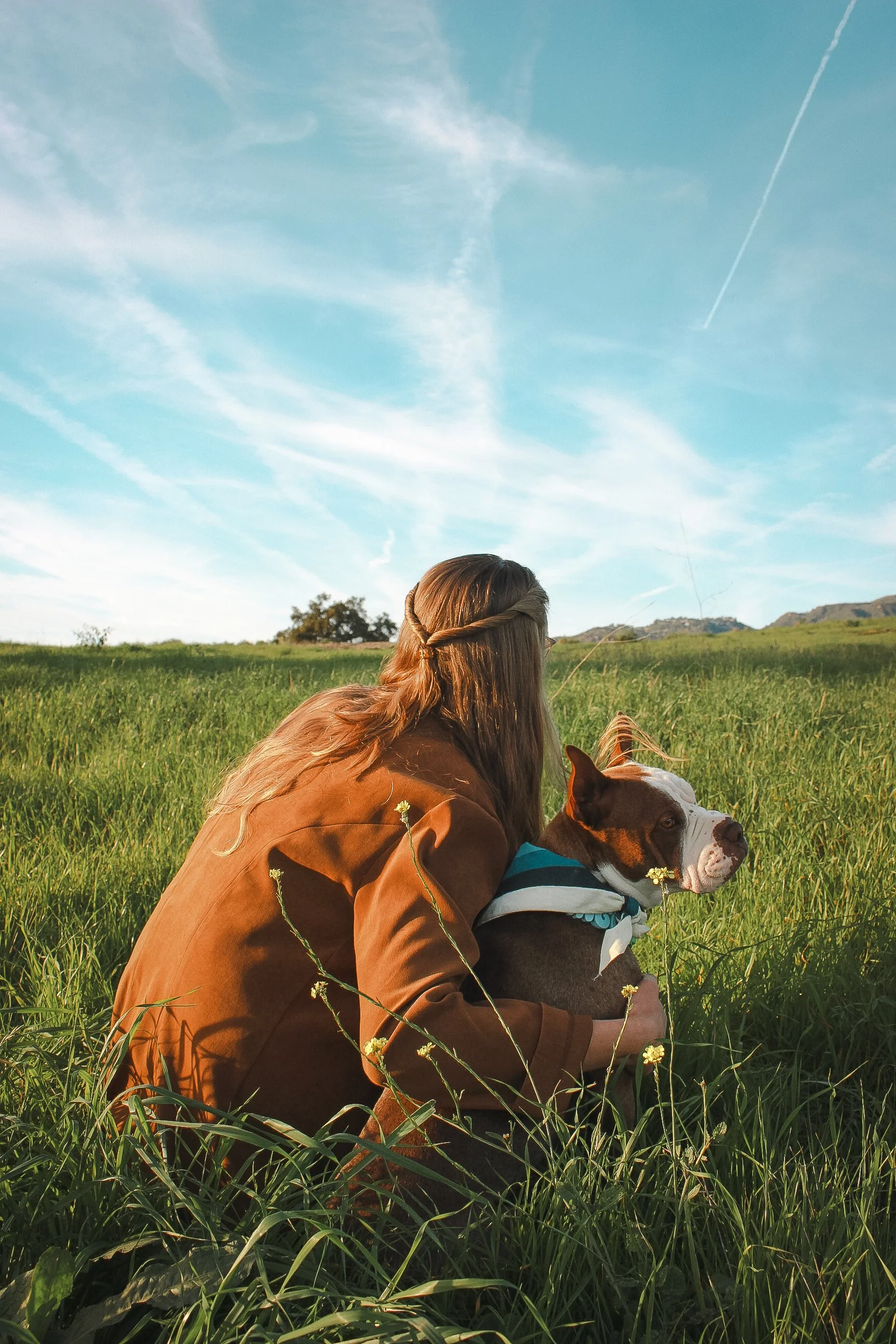 A woman with long hair and a brown jacket sitting in a grassy field with a dog in Montecito