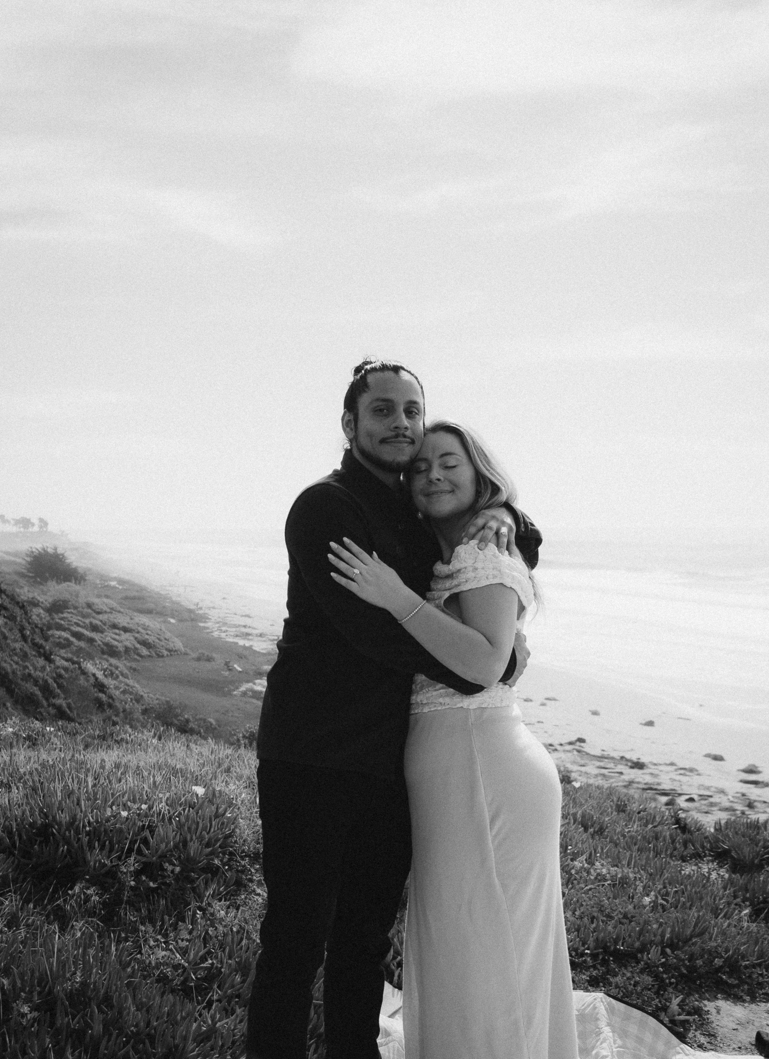 A couple embracing on a grassy cliff near the ocean, smiling and hugging each other, with the beach and water in the background.