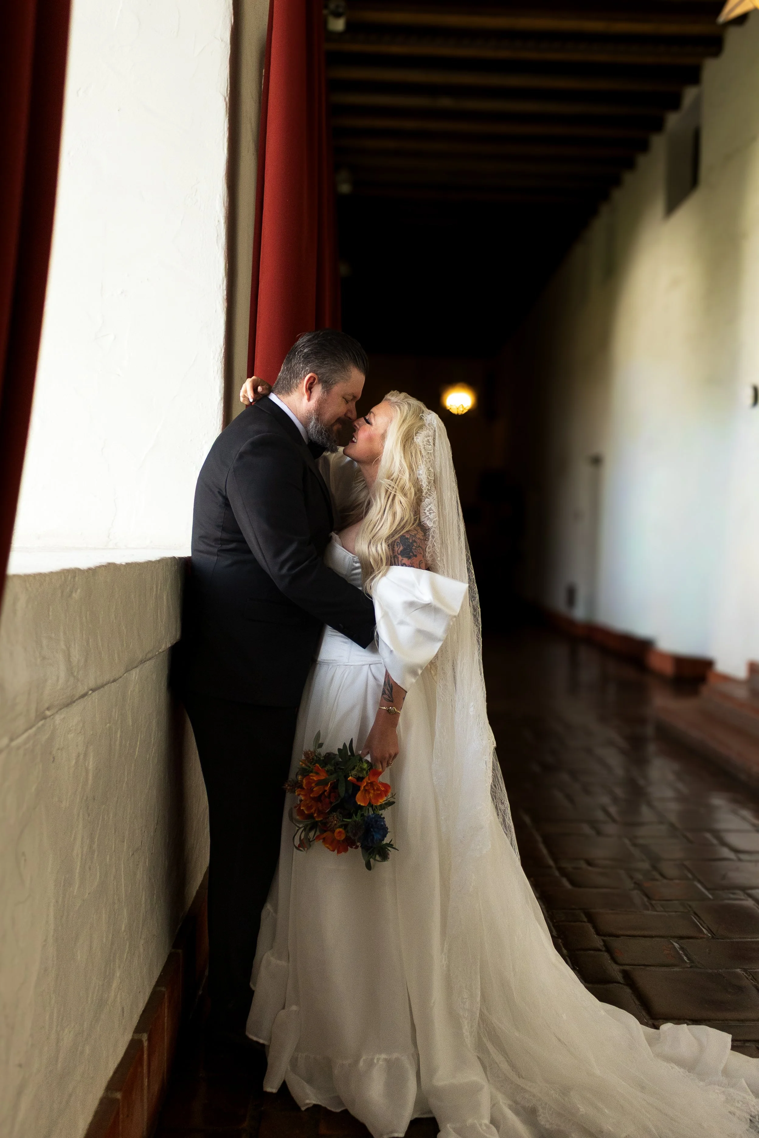 A couple in wedding attire sharing a tender moment indoors, with the bride holding a bouquet of flowers and leaning towards the groom.