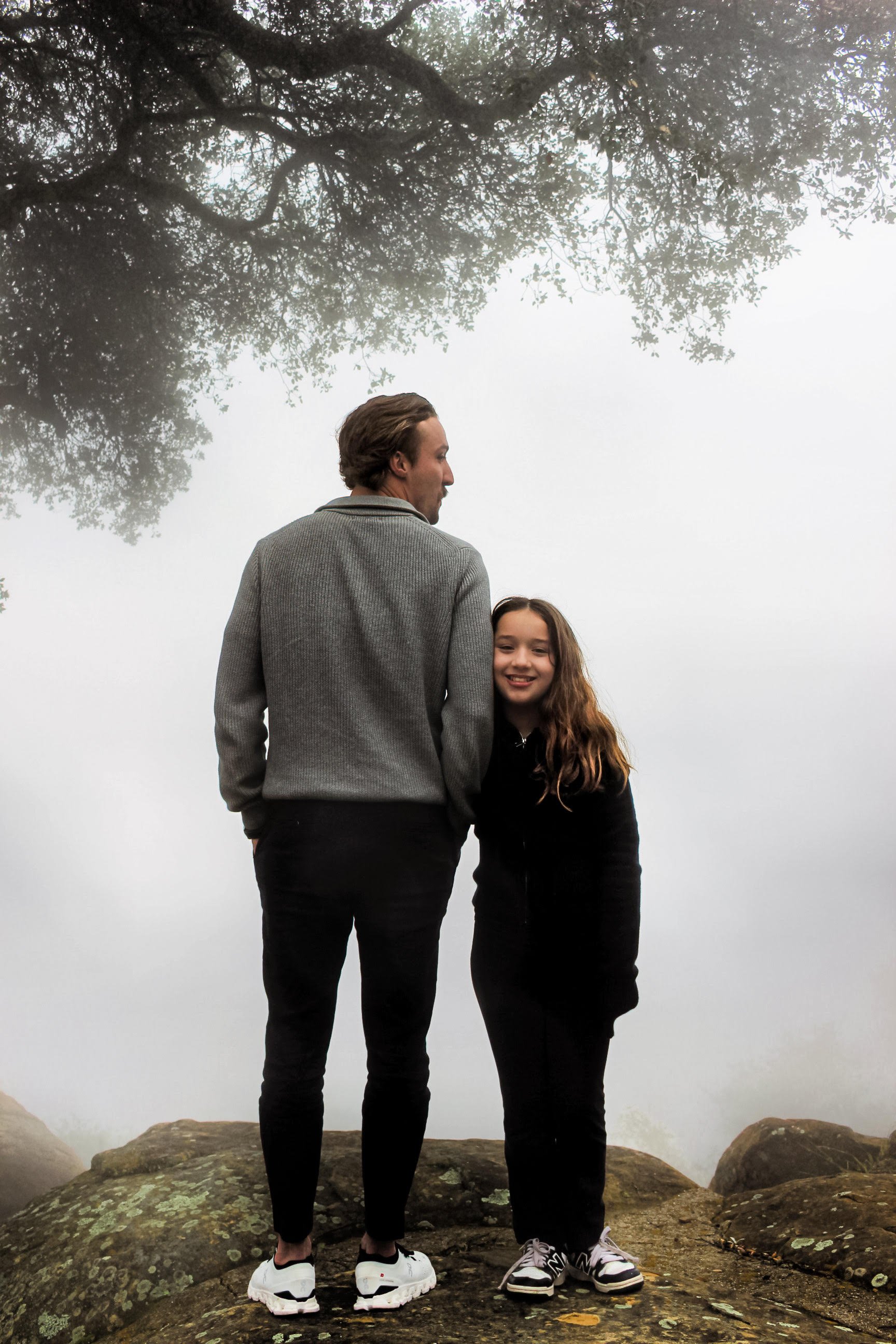 A man and a girl standing on rocks outdoors with fog and a large tree overhead in the background.