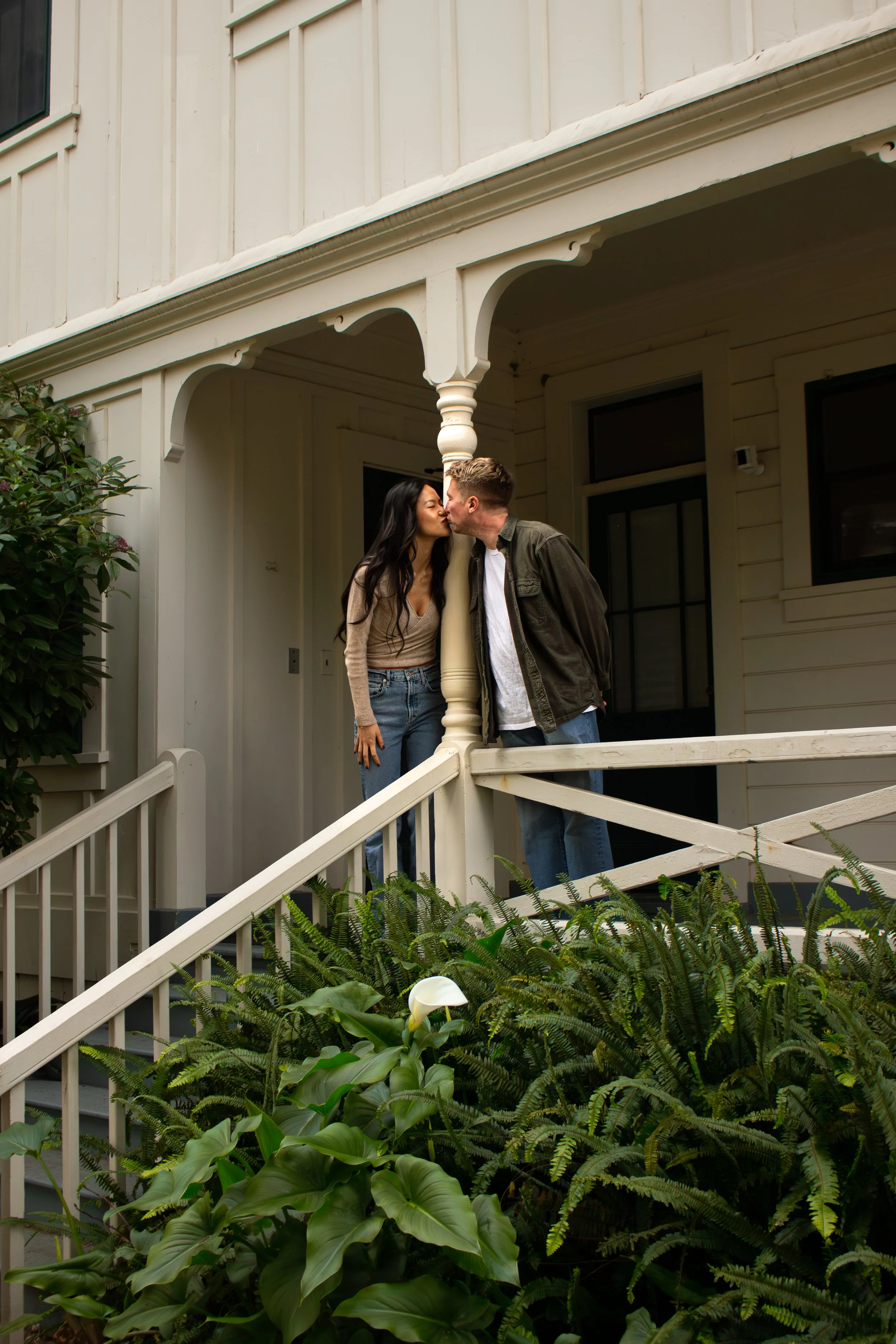 A couple sharing a kiss on a porch of a house surrounded by green plants and flowers in SB