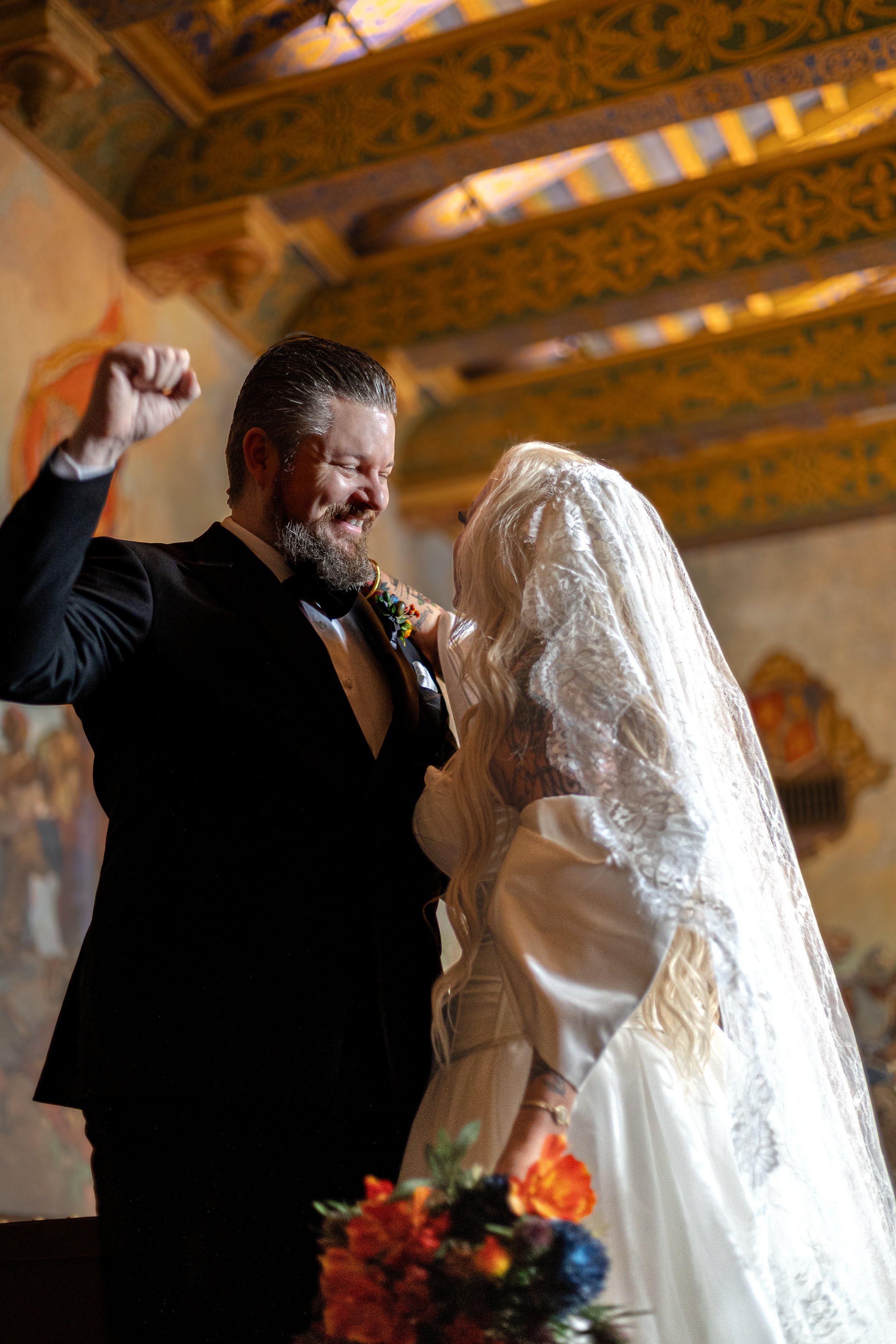 A wedding couple sharing a joyful moment, the man in a black tuxedo with a beard and the woman in a white lace wedding dress with long hair and a veil, holding a colorful bouquet, inside a decorated venue with ornate woodwork on the ceiling.