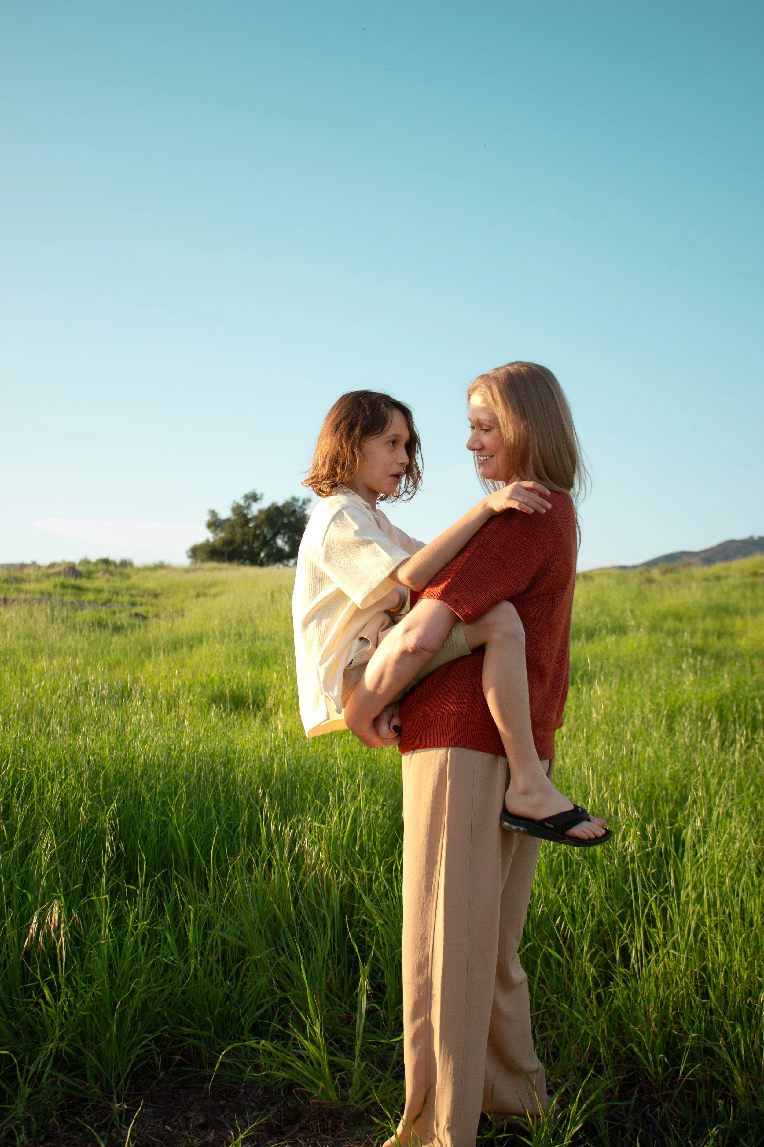 A woman with blonde hair wearing a red shirt is holding a young girl with short brown hair. The girl is wearing a beige shirt and shorts, and she is sitting on the woman's hip. They are outdoors in a green field with hills in the background under a c