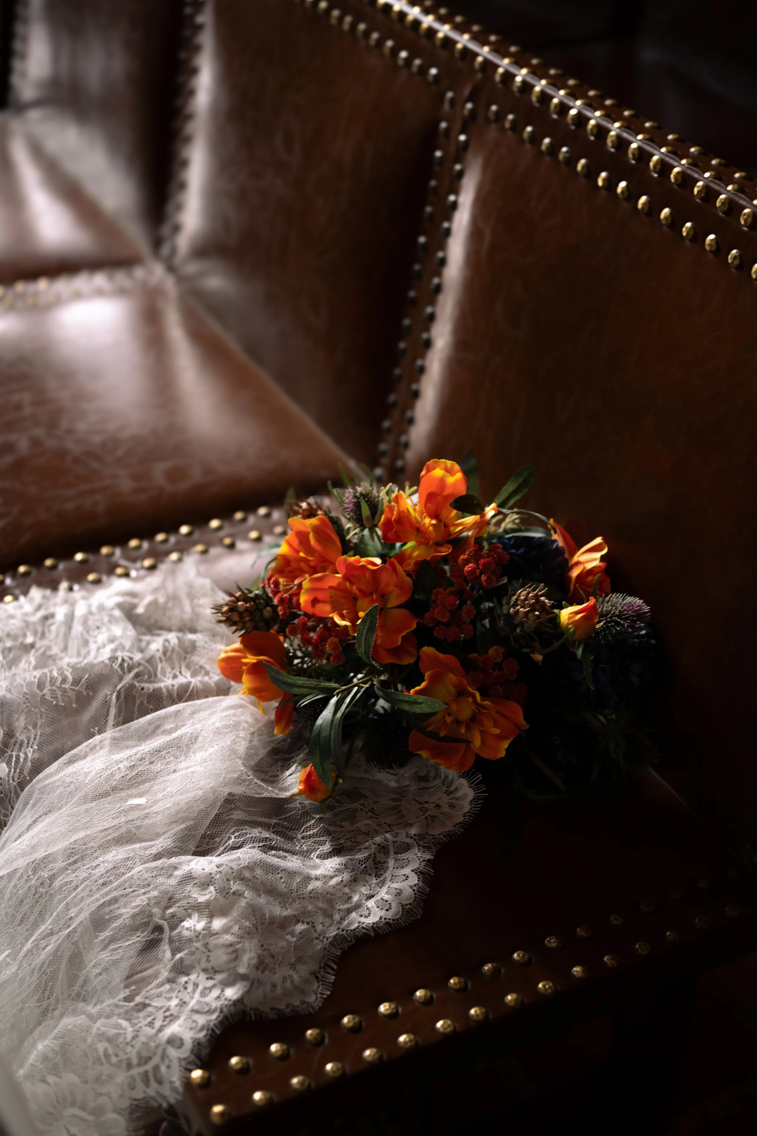 A bouquet of orange, red, and purple flowers resting on a antique leather sofa with detailed brass nailhead trim, with a lace cloth partly draped over the seat.