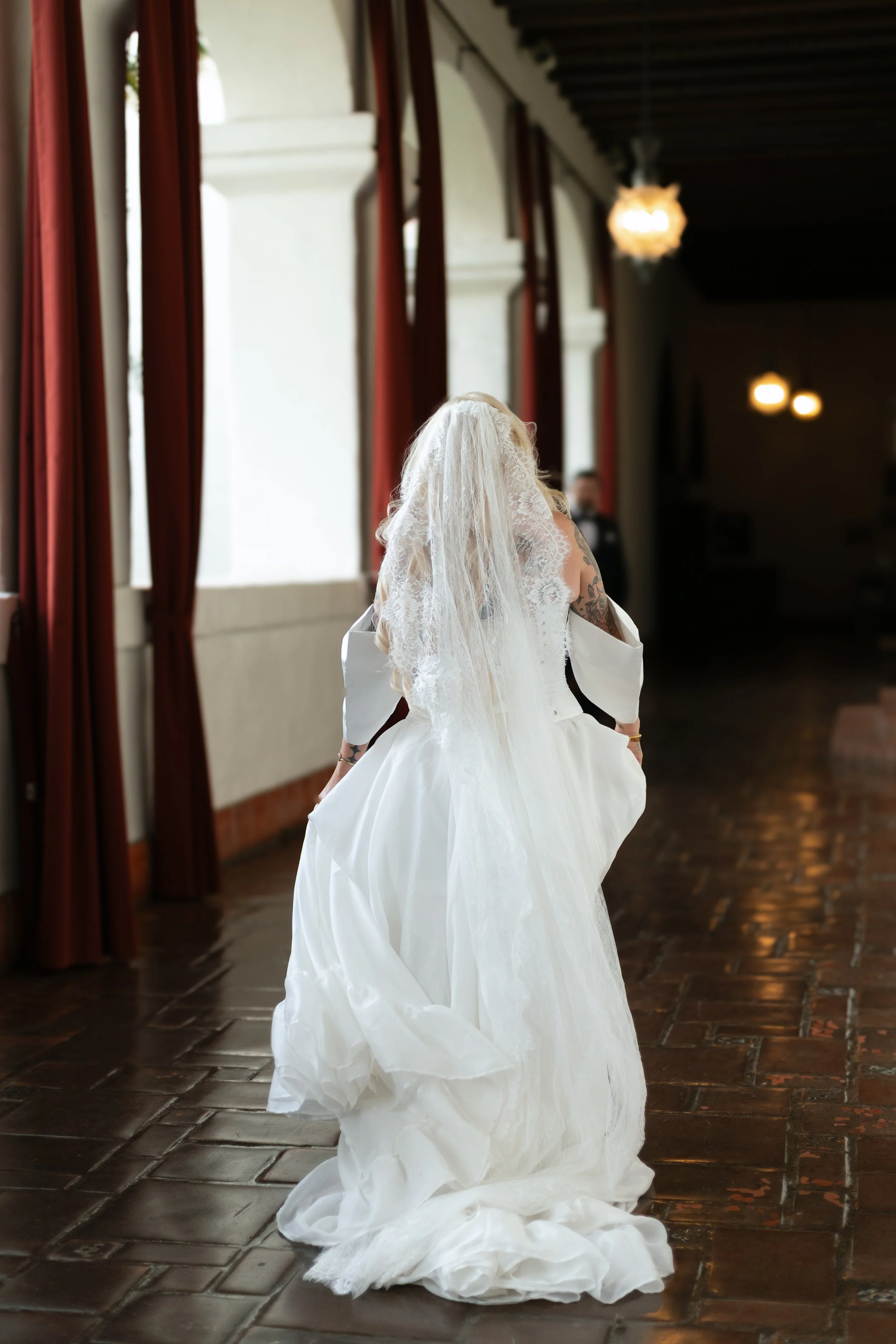 A bride in a white wedding dress and veil walking down a hallway with large windows and red curtains.