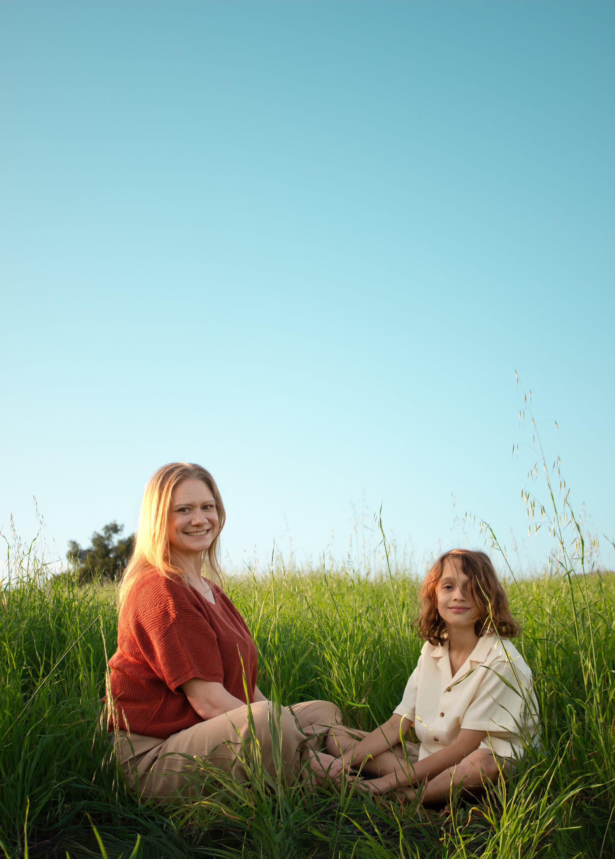 A woman and her son sitting cross-legged in a grassy field under a clear blue sky, smiling at the camera.