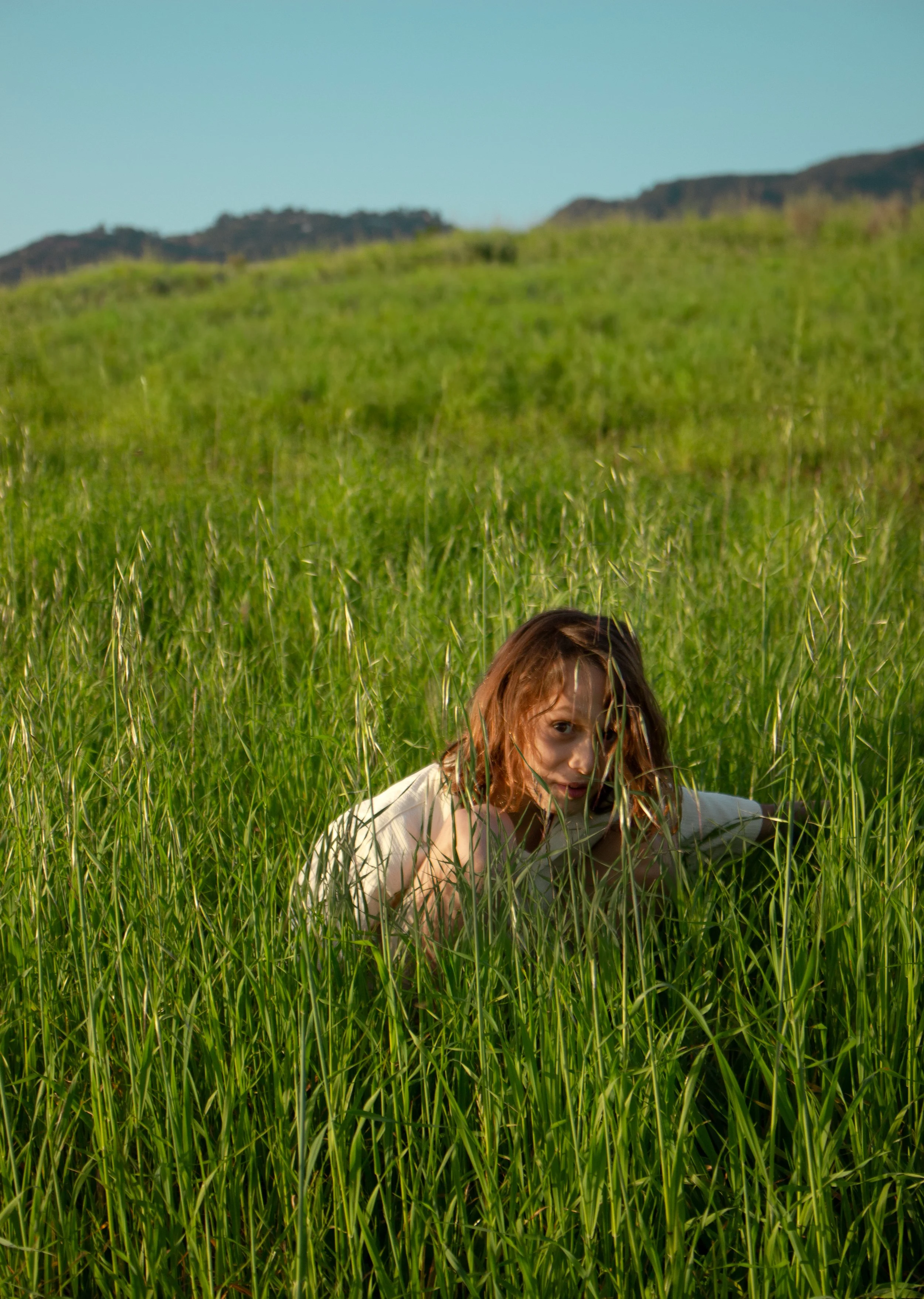 A boy with reddish-brown hair crawling through a tall, green grassy field with hills and a blue sky in the background.