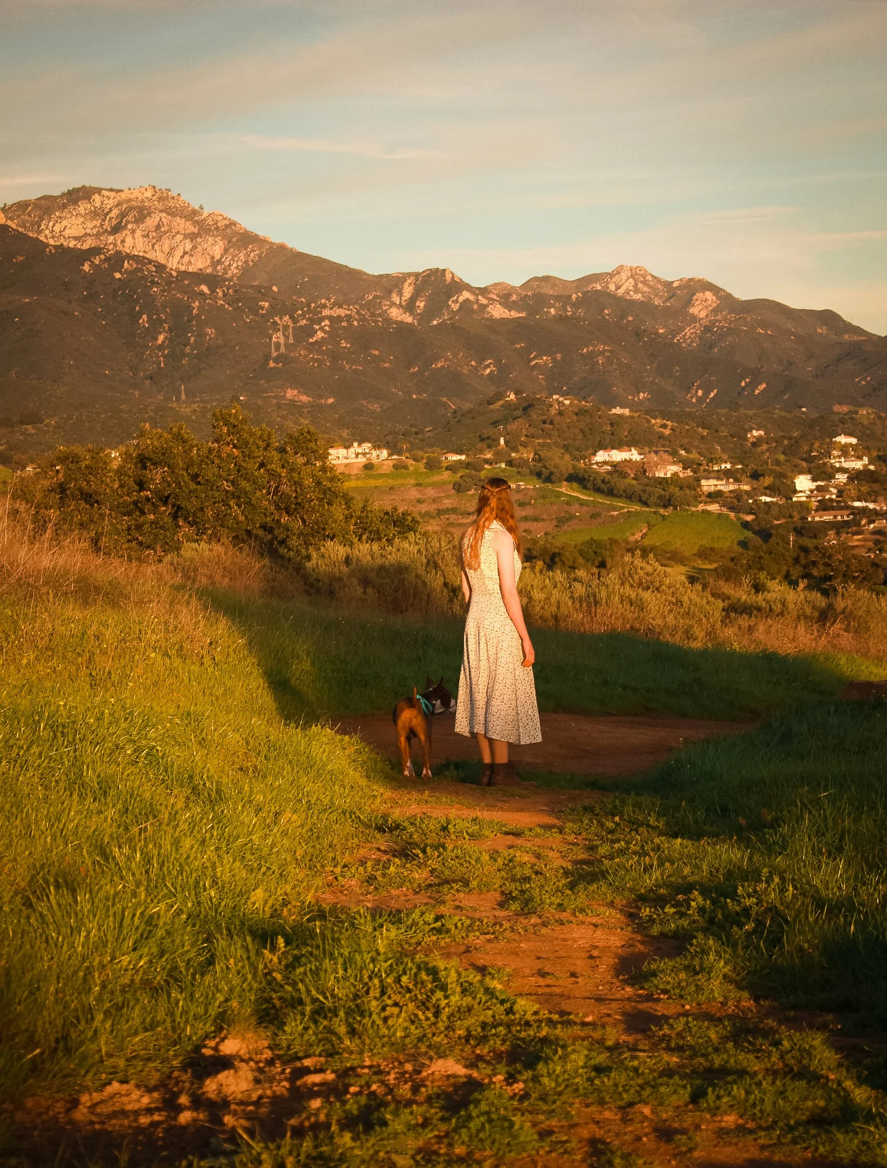 A woman in a floral dress walking with a dog on a leash along a dirt trail in a grassy field during golden hour with mountains in the background.