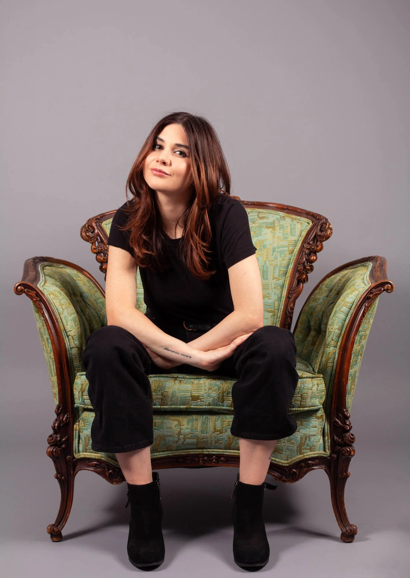 A young woman with long brown hair and wearing a black t-shirt, black pants, and black boots sits on an ornate vintage green and gold upholstered chair against a plain gray background.