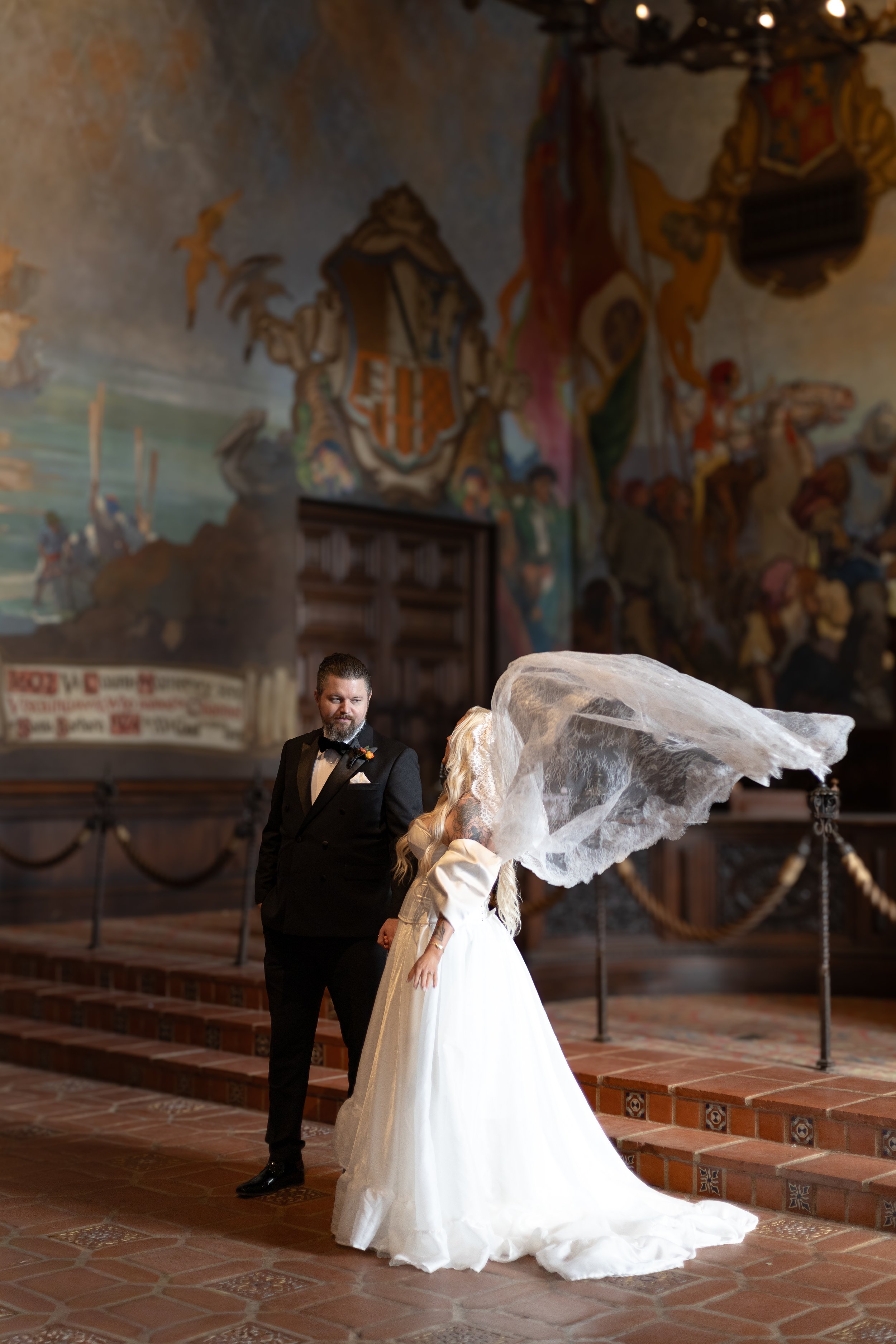 A bride and groom standing in the Santa Barbara courthouse, with the bride gently bowing her head as her veil blows in the wind, while the groom looks at her. The hall has ornate murals and a wooden door in the background.